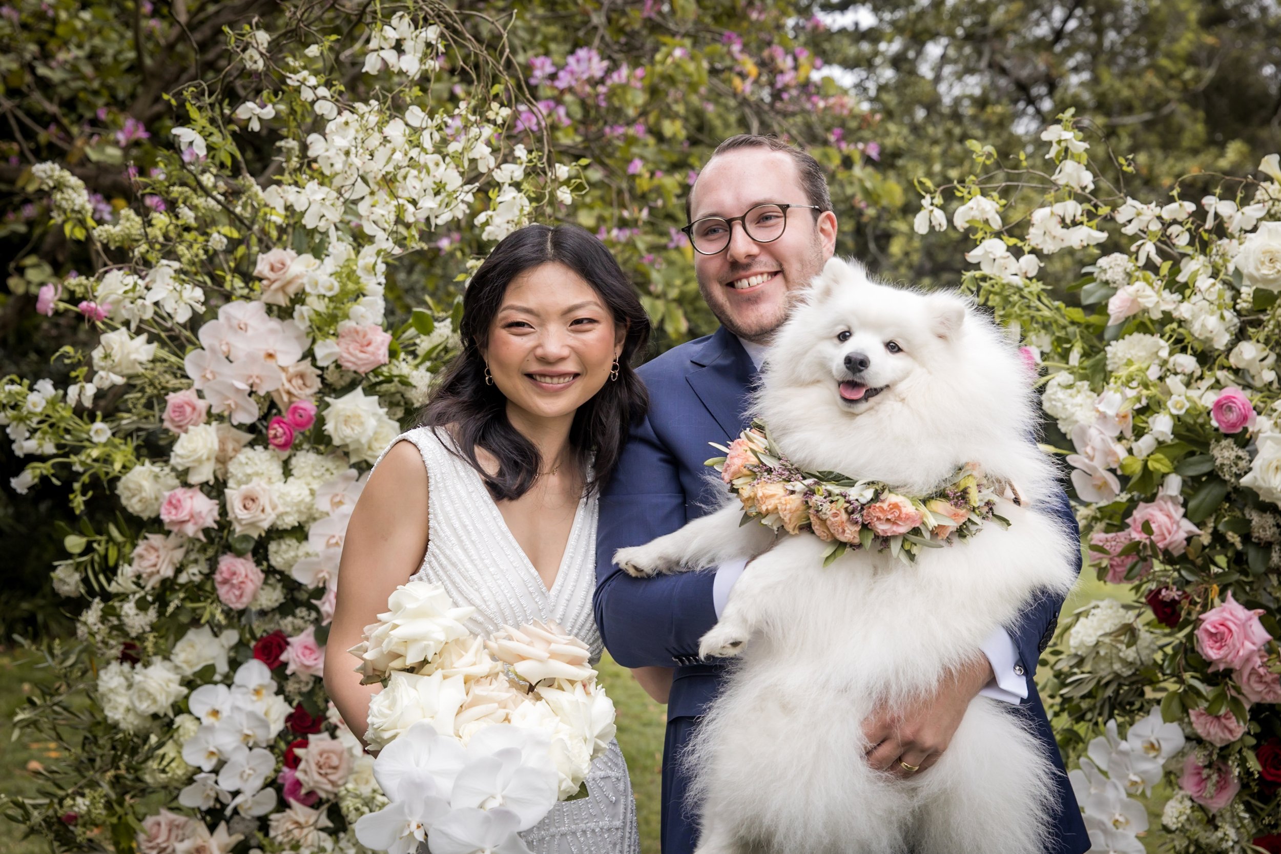A newlywed couple standing outdoors surrounded by a floral arch. The woman is holding a bouquet of white roses, the man is holding a fluffy white dog wearing a floral wreath. Vaucluse House Sydney - Katsu Nojiri Photographer