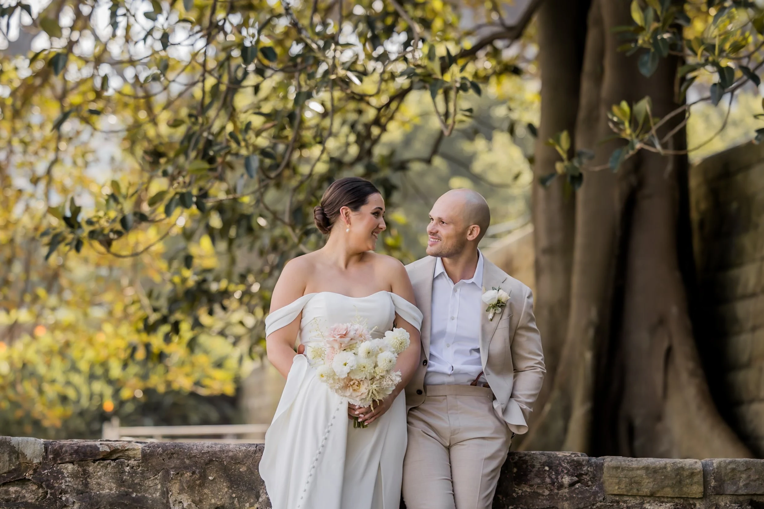 A newlywed couple, a woman in a white wedding dress holding a bouquet and a man in a light-colored suit, sitting on a stone wall outdoors with trees in the background, sharing a happy moment.