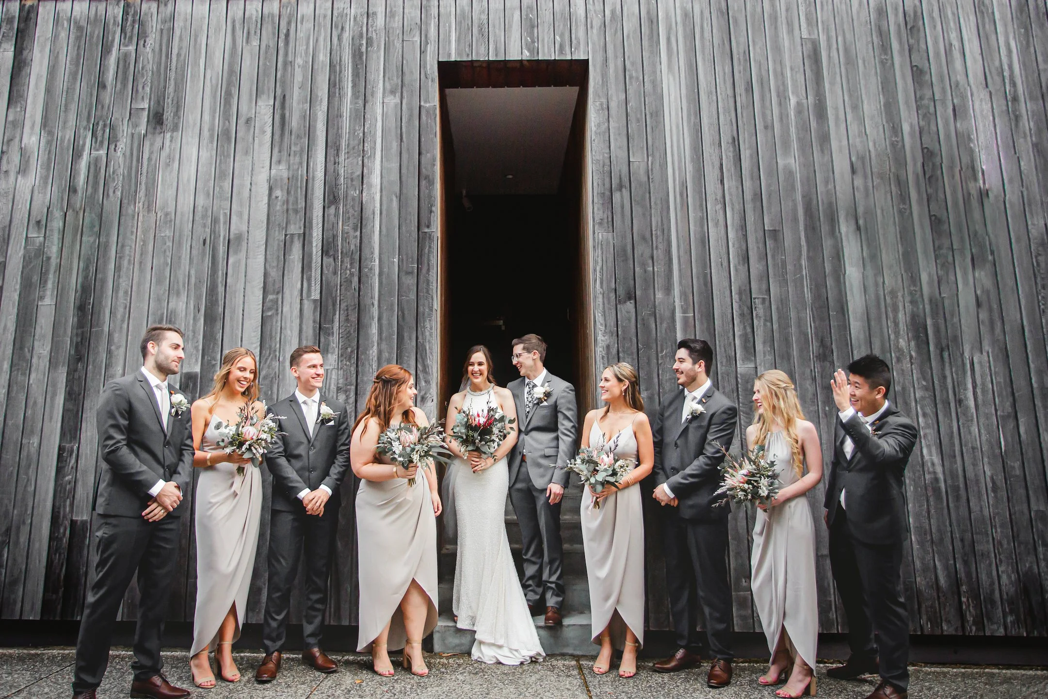 A wedding party standing in front of a modern dark wooden wall, posing for a photo. The group includes the bride and groom in the center, surrounded by bridesmaids and groomsmen holding bouquets - Sergeants Mess Sydney - Katsu Nojiri Photographer