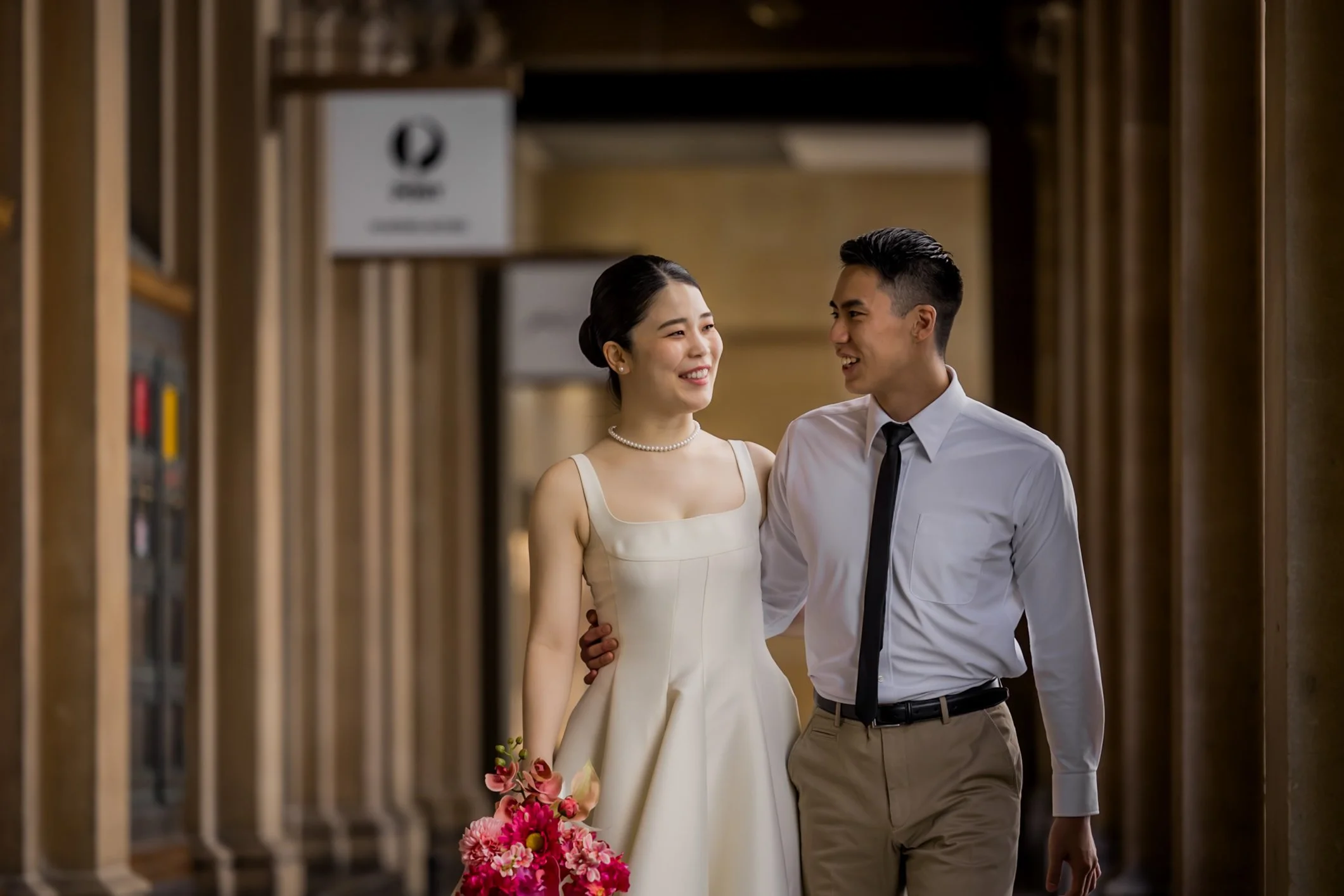 A smiling woman in a white dress and pearl necklace holding a bouquet of pink and red flowers, walking arm-in-arm with a man in a white shirt and black tie in a hallway.