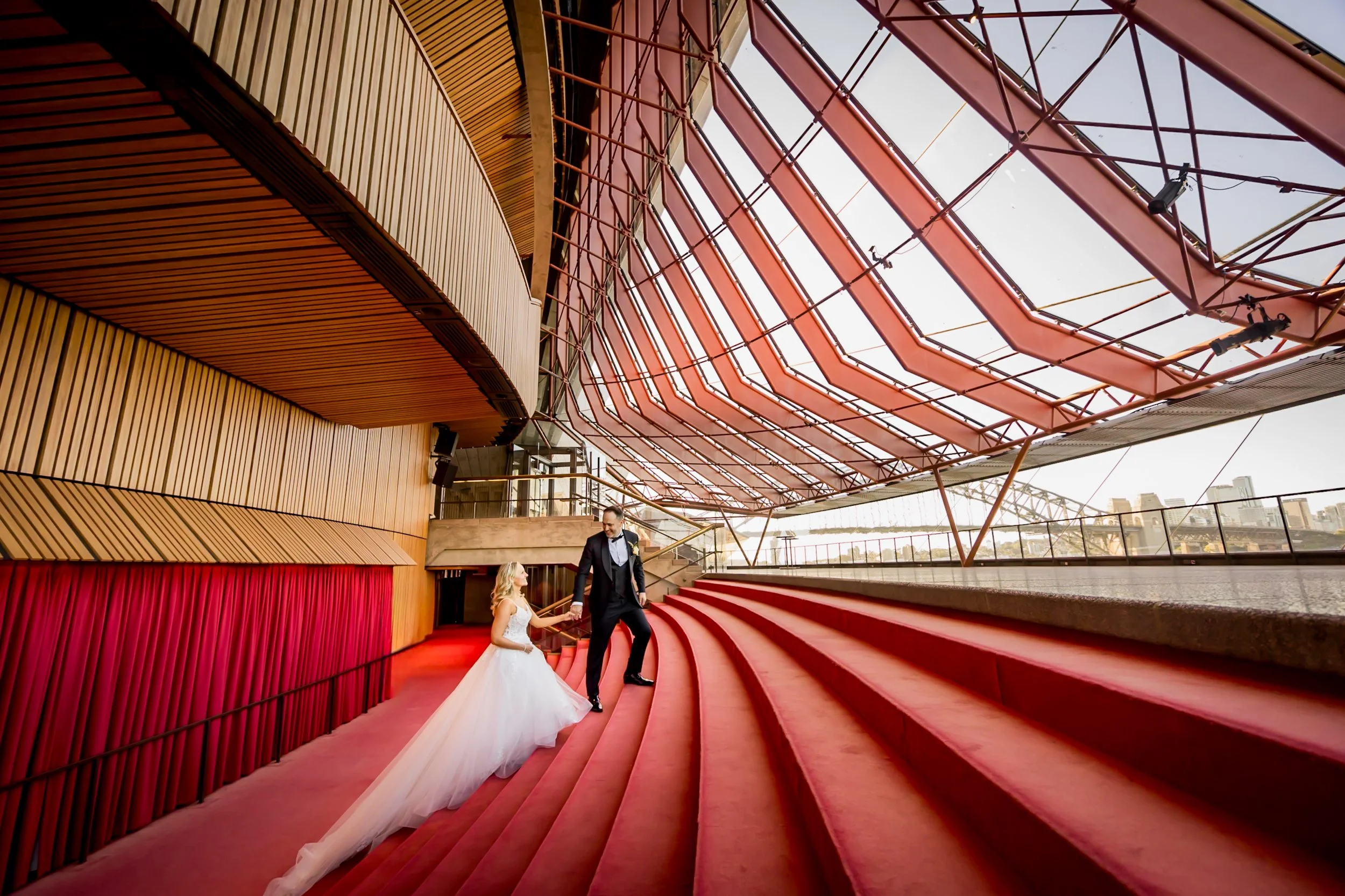 A bride and groom on red carpeted stairs inside Sydney Opera House with a glass and metal roof structure.