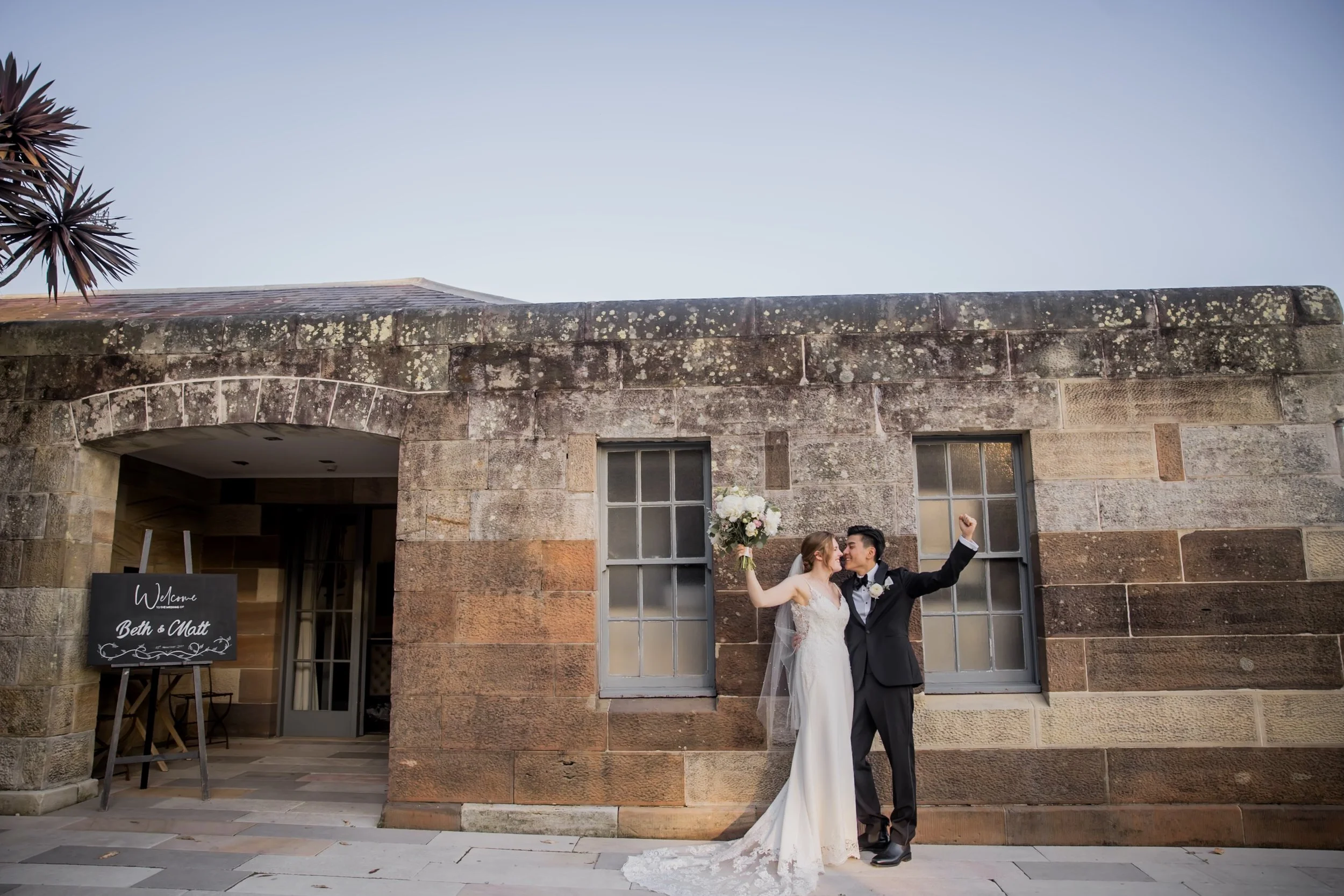 A newlywed couple in wedding attire celebrating in front of a stone building, with the bride holding a bouquet and the groom raising a fist, as a sign welcoming guests to a wedding event.