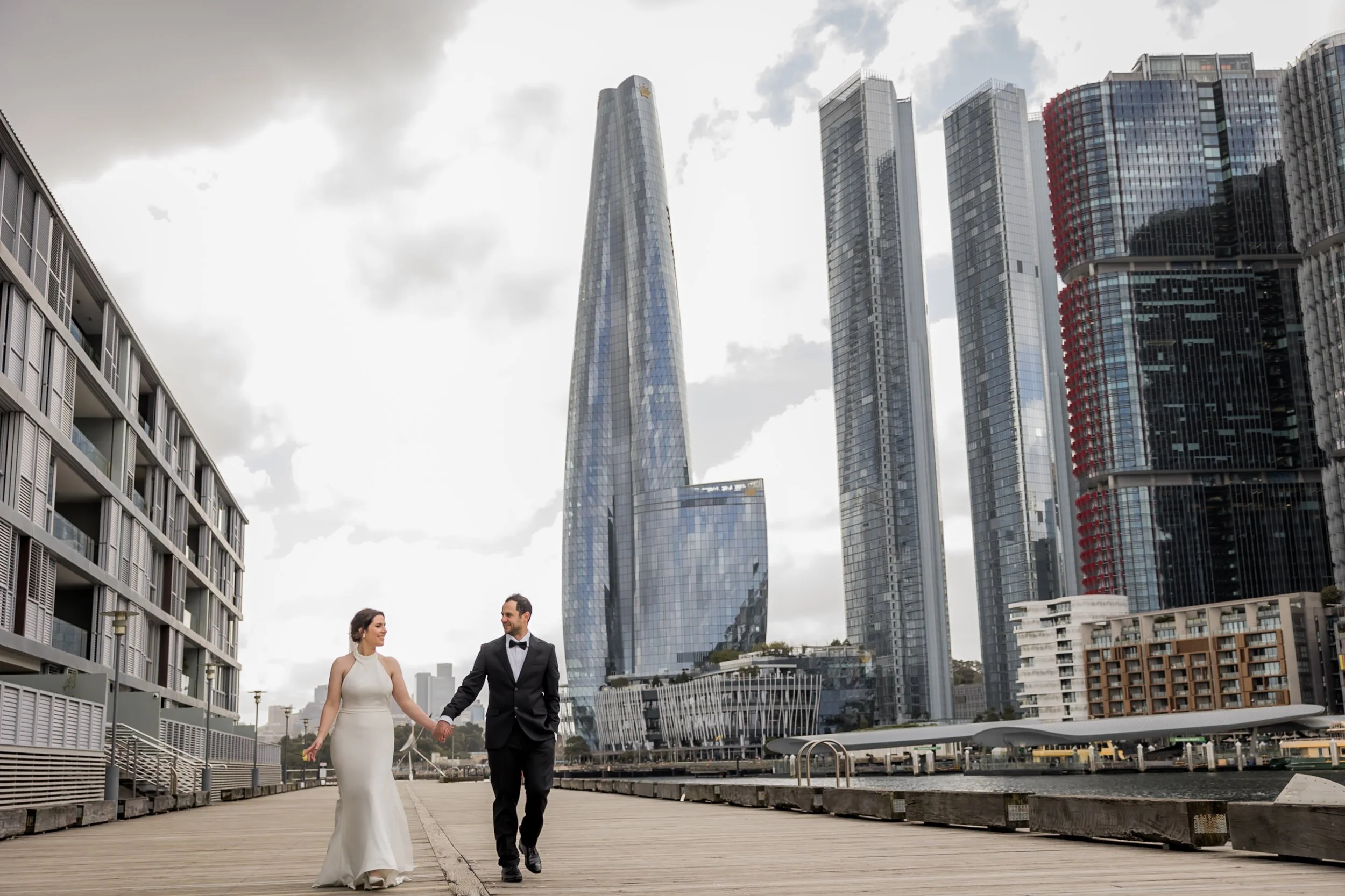 A bride and groom holding hands and walking on a wooden pier in front of modern skyscrapers and high-rise buildings in a city