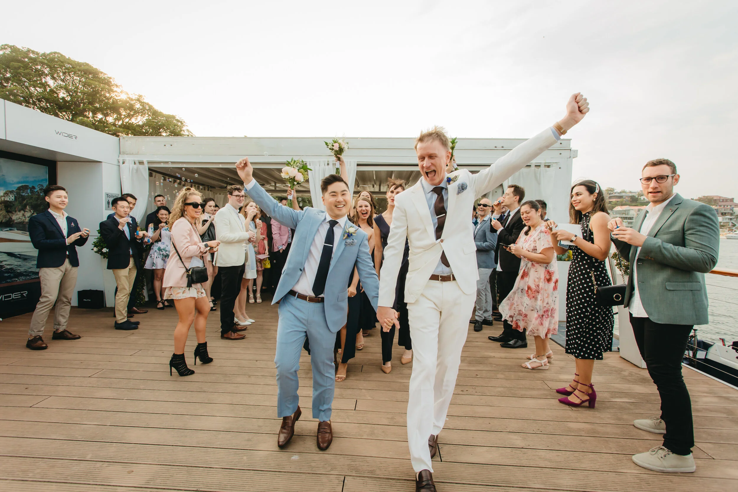 Two men in suits holding hands and celebrating at a wedding reception on a wooden deck near the water, surrounded by guests clapping and smiling.