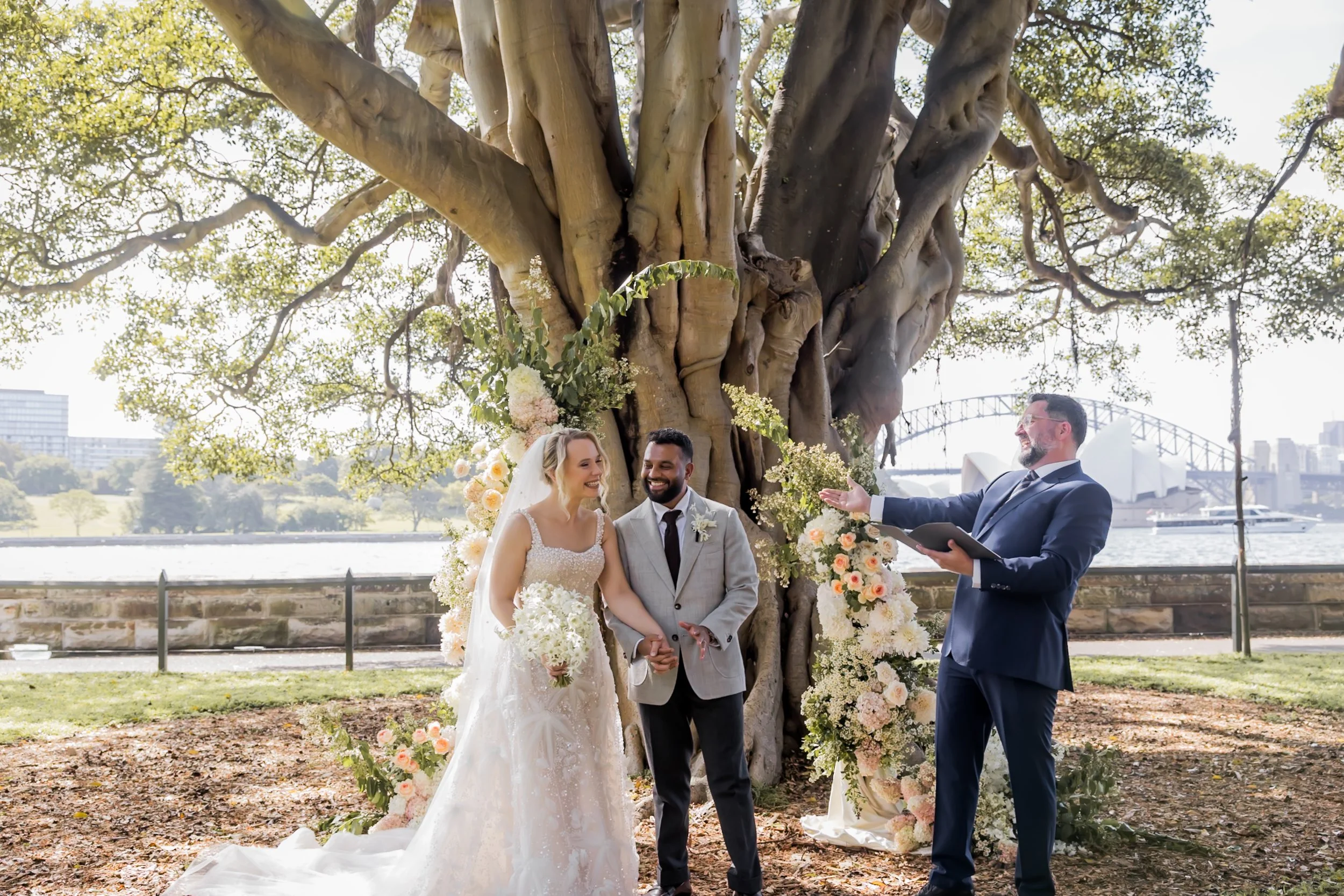A couple getting married outdoors next to a large tree with flowering decorations, officiant speaking to them by a river with cityscape in the background.  Botanic House Sydney - Katsu Nojiri Sydney Wedding Photographer