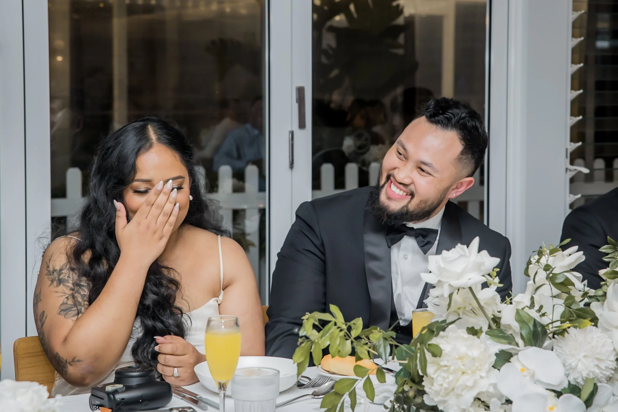 A woman with long dark hair and a tattoo on her arm is covering her mouth and laughing, while a man in a tuxedo with a bow tie is smiling at her at a wedding reception. There are flowers, drinks, and table settings in front of them.