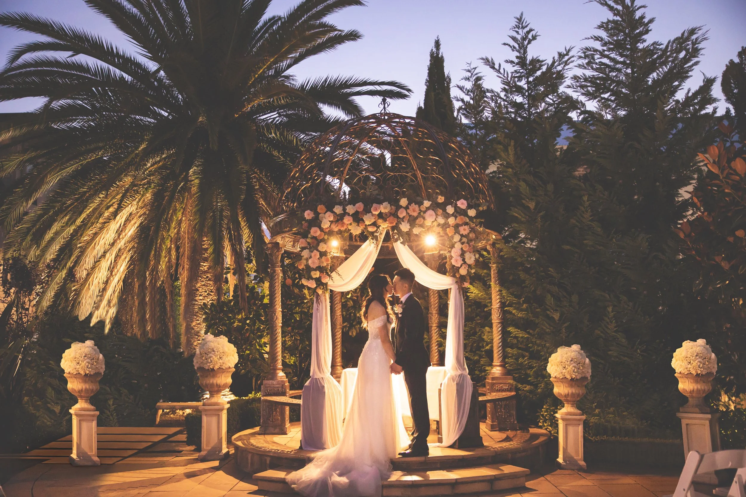 A bride and groom standing face-to-face and holding hands under a wedding arch adorned with flowers and drapes, surrounded by lush plants and trees during the evening outdoor wedding ceremony. - Lauriston House