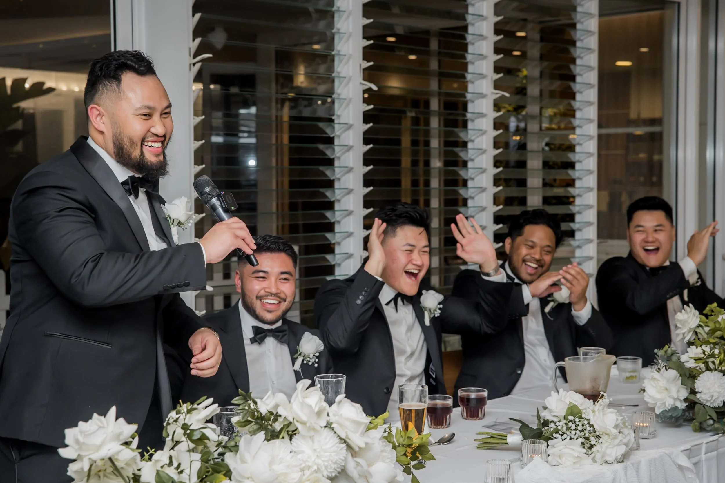 Groom giving a speech at a wedding reception, surrounded by groomsmen laughing and smiling, with floral decorations and drinks on the table. Long Reef Golf Course  Sydney - Katsu Nojiri Sydney Wedding Photographer