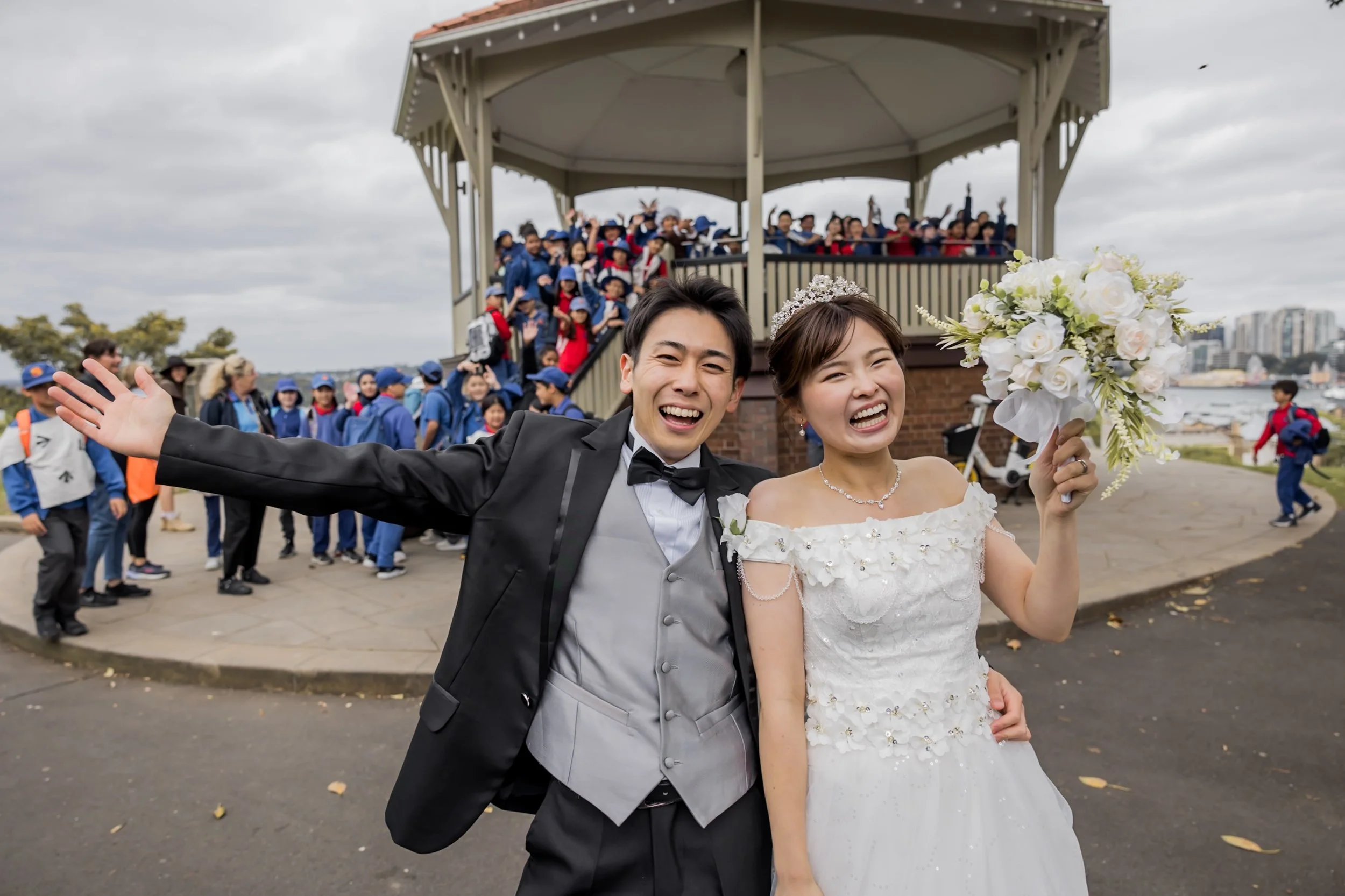 A newlywed couple, the groom in a tuxedo and the bride in a wedding dress, celebrate outdoors with smiles, flowers, and a large group of children and teenagers in the background at a park.