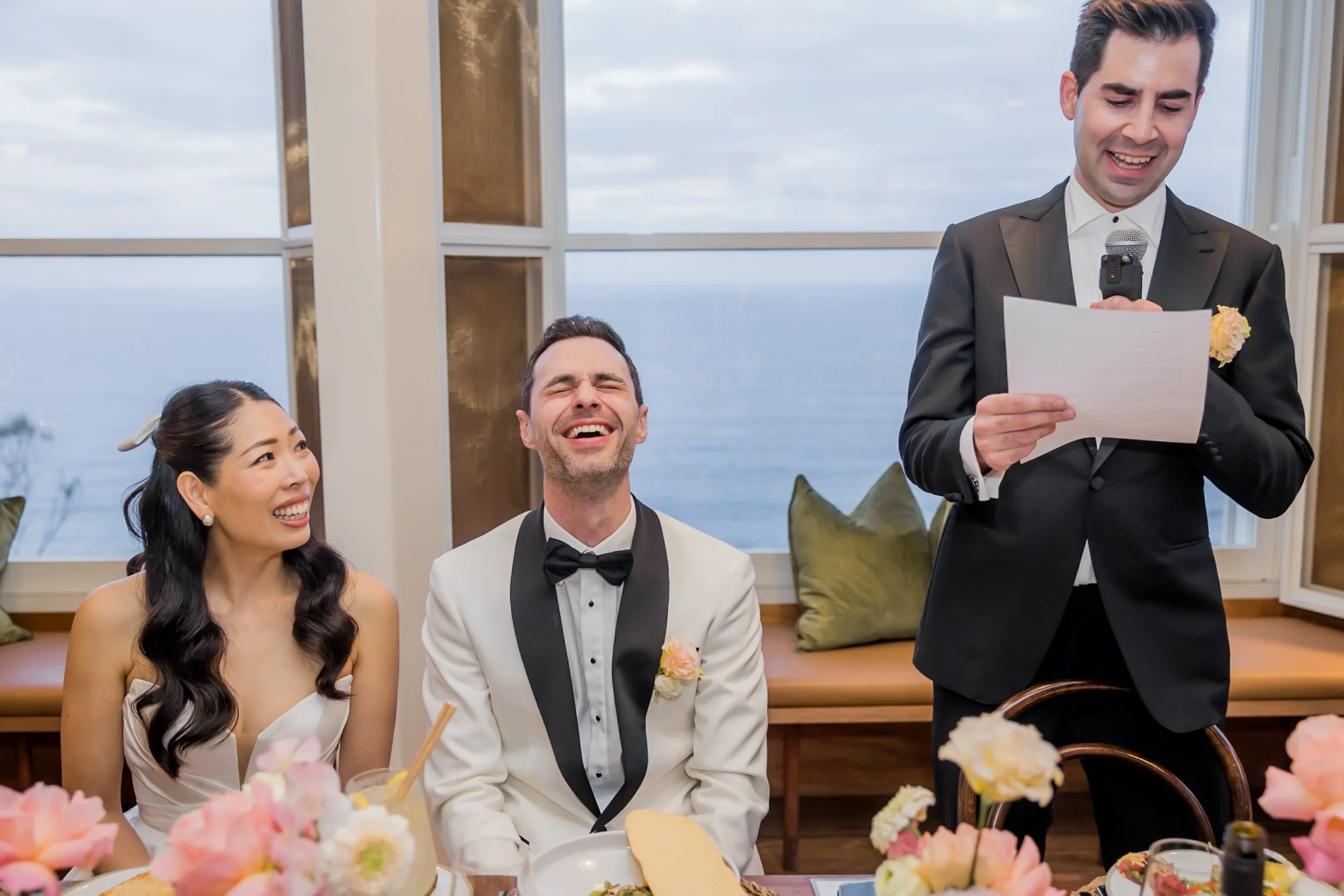 A wedding reception with a woman and a man sitting at a table, laughing, while a man in a black tuxedo is giving a speech with a microphone in front of a window overlooking the ocean.