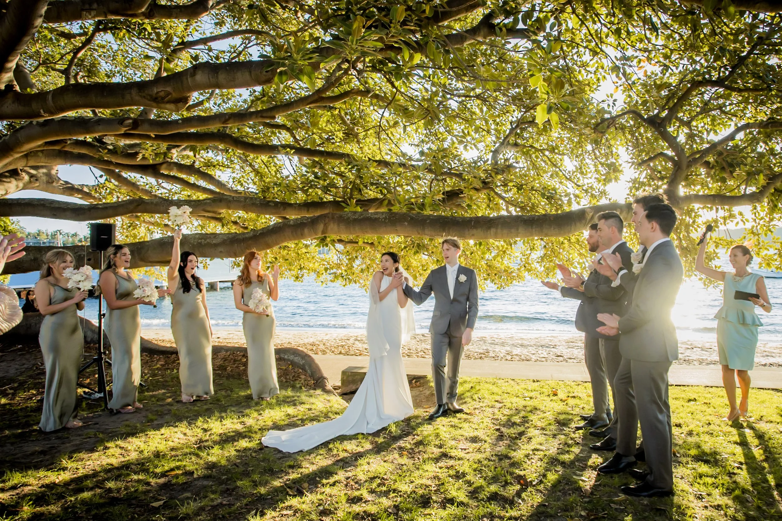 A wedding ceremony takes place under a large tree on a beachside, with a bride and groom in the center surrounded by bridesmaids and groomsmen, holding bouquets, clapping, and celebrating during sunset.