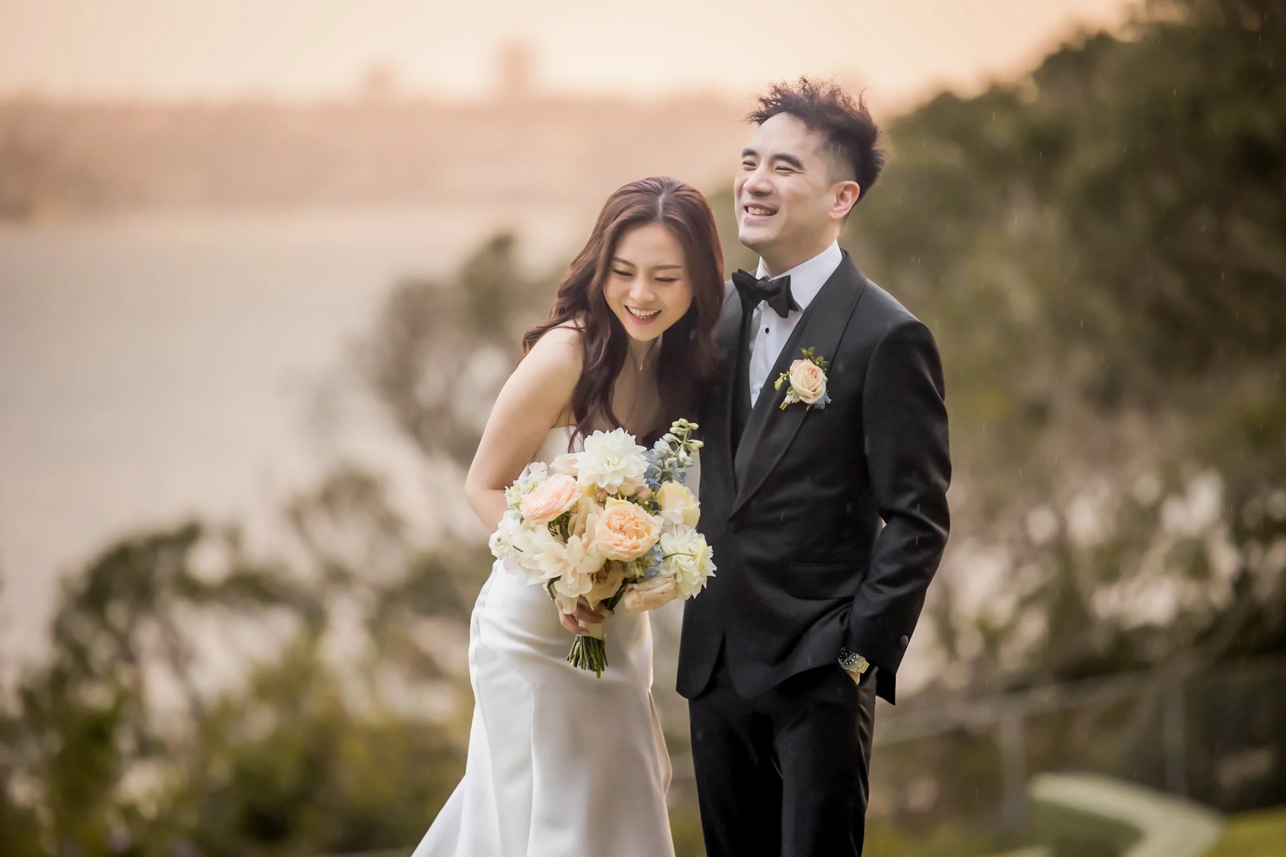 A bride and groom in wedding attire smiling outdoors during sunset, with the bride holding a bouquet of flowers - Sergeants Mess