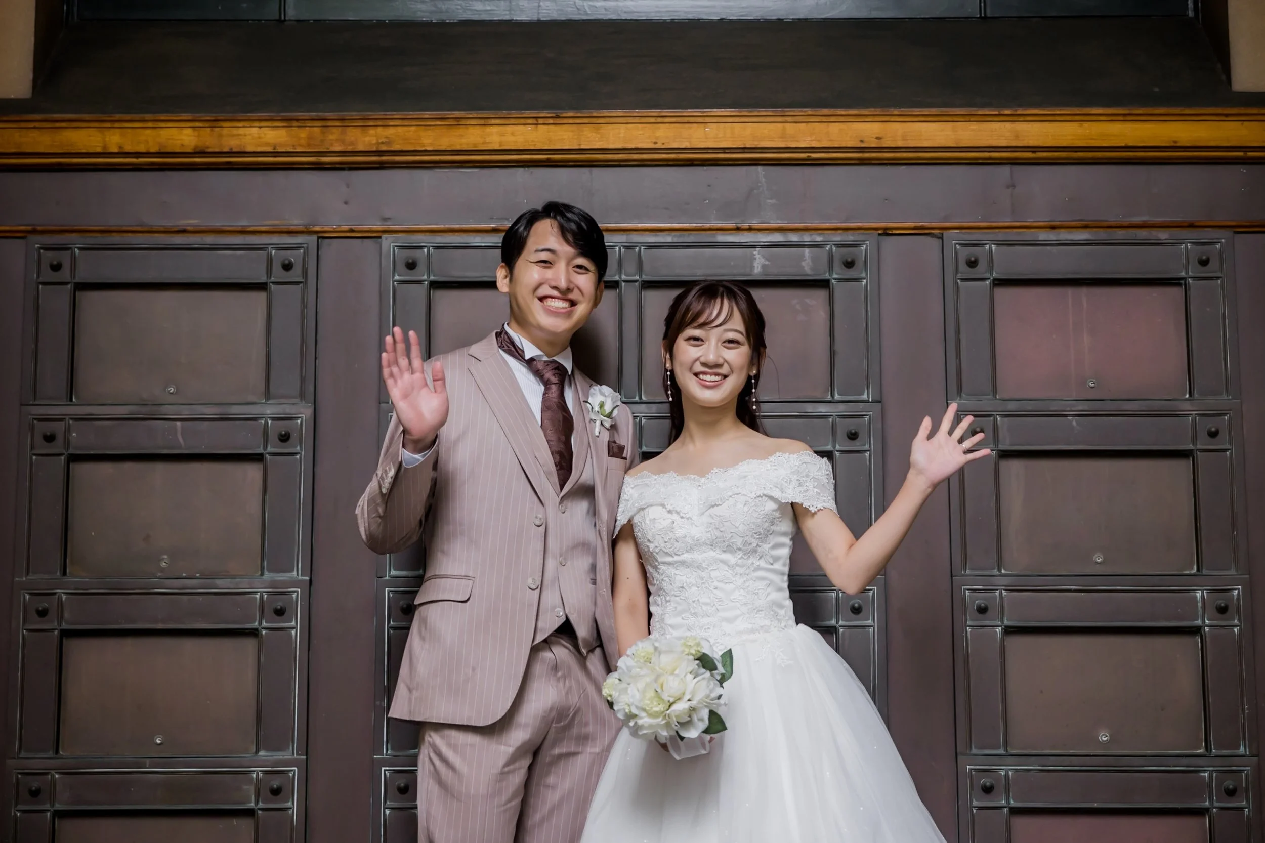 A smiling bride and groom waving at the camera, standing in front of a metal wall with wooden accents, at a wedding celebration.