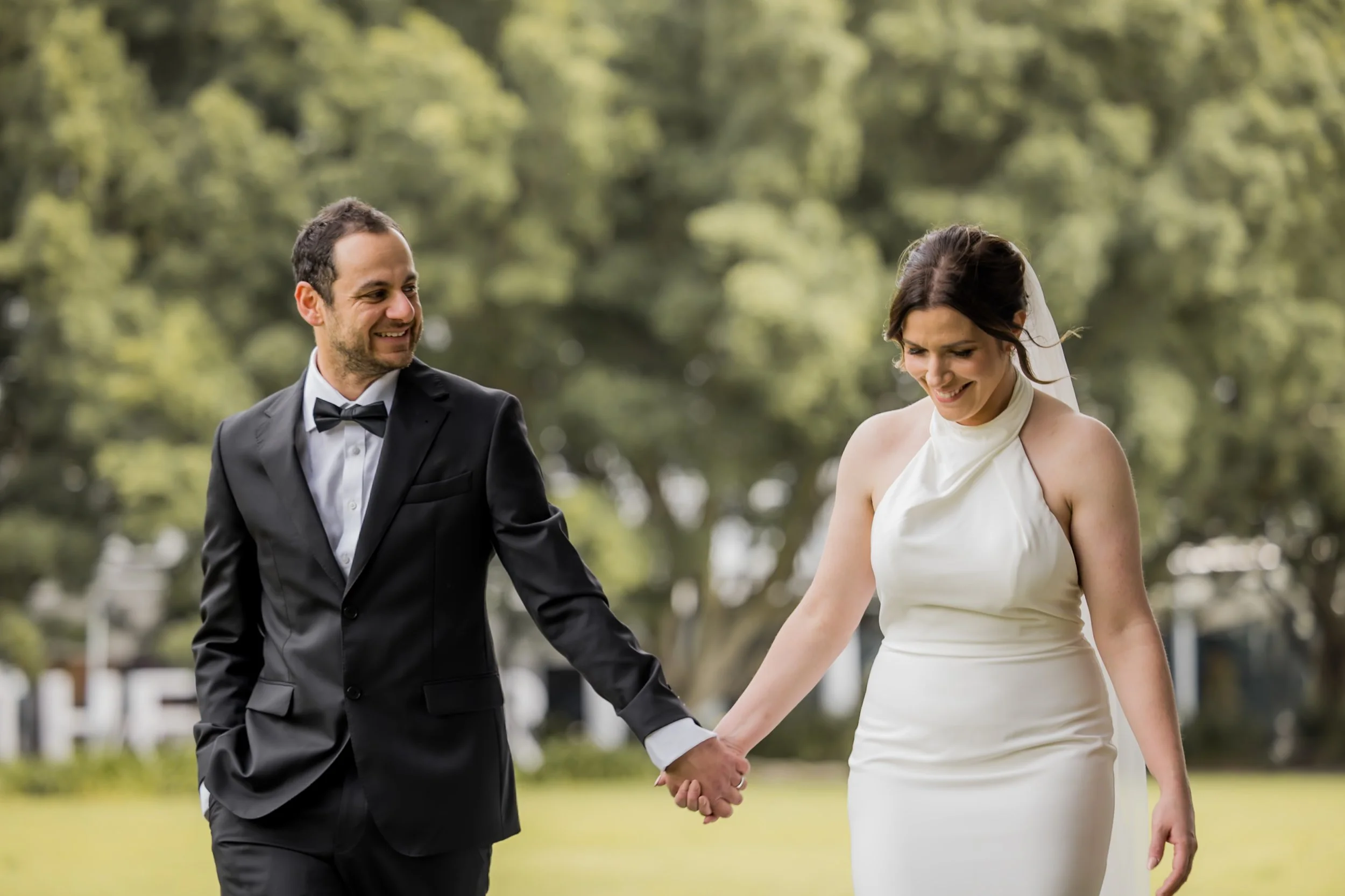 A bride and groom walking hand in hand during their outdoor wedding, surrounded by trees and greenery.