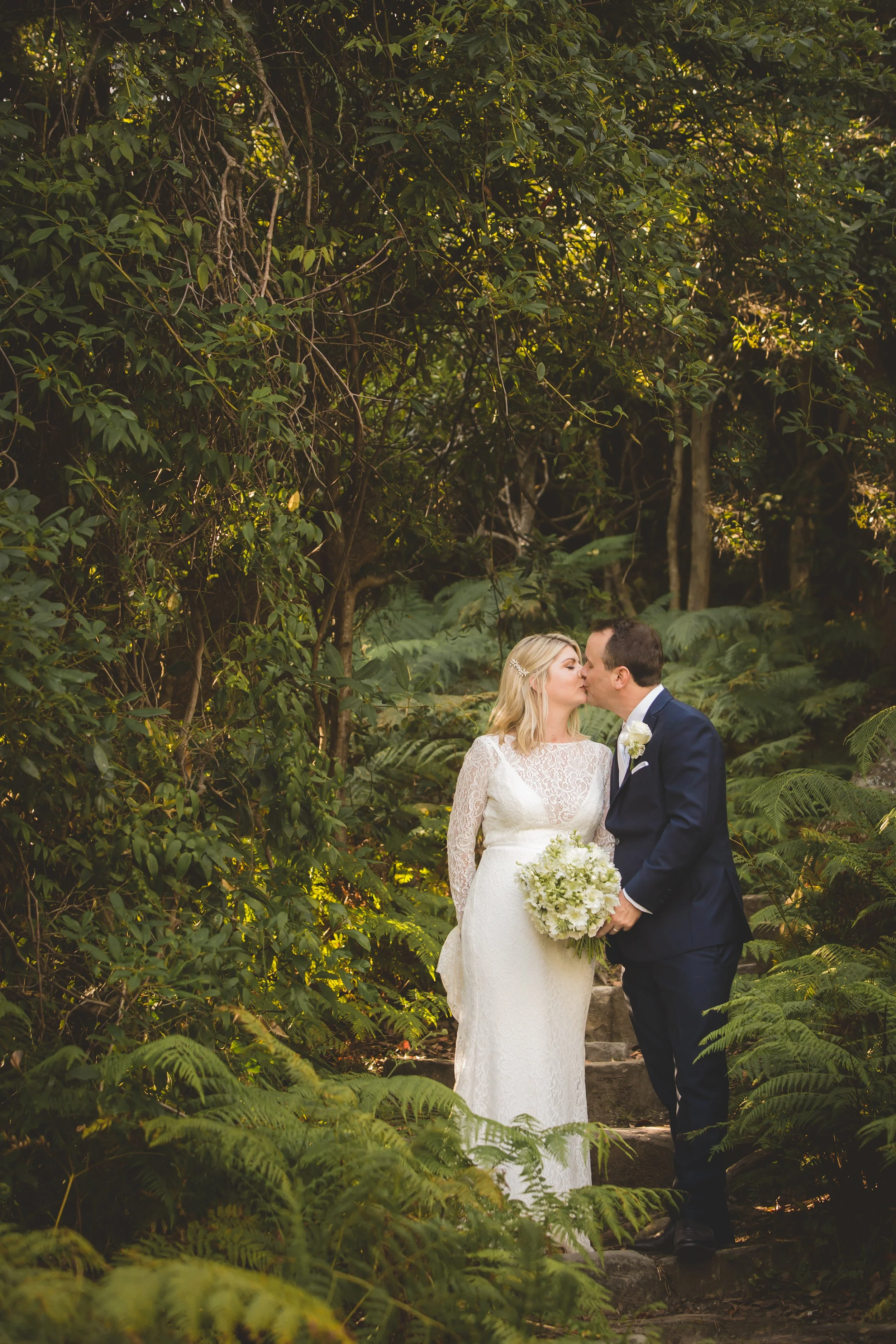 A bride and groom kissing in a lush forest setting, with greenery and ferns surrounding them at Sergeants Mess