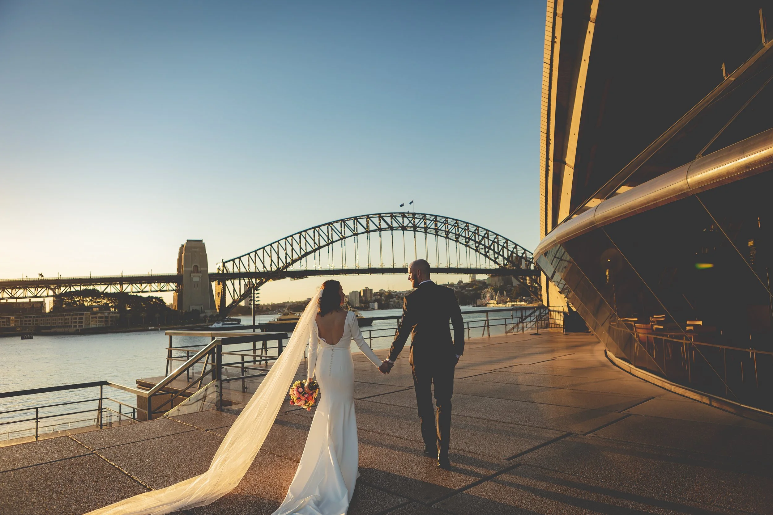 A bride and groom walking hand in hand along a waterfront promenade during sunset, with the Sydney Harbour Bridge visible in the background.