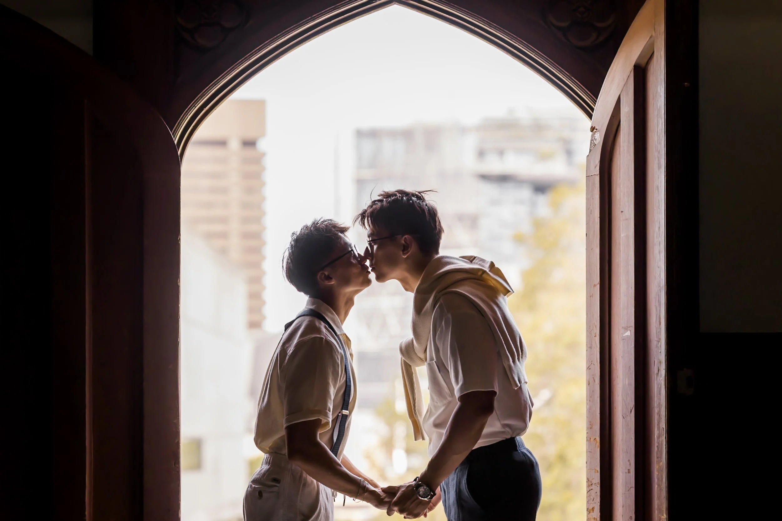 A couple stands in an arched doorway, about to kiss, holding hands, with city buildings in the background.