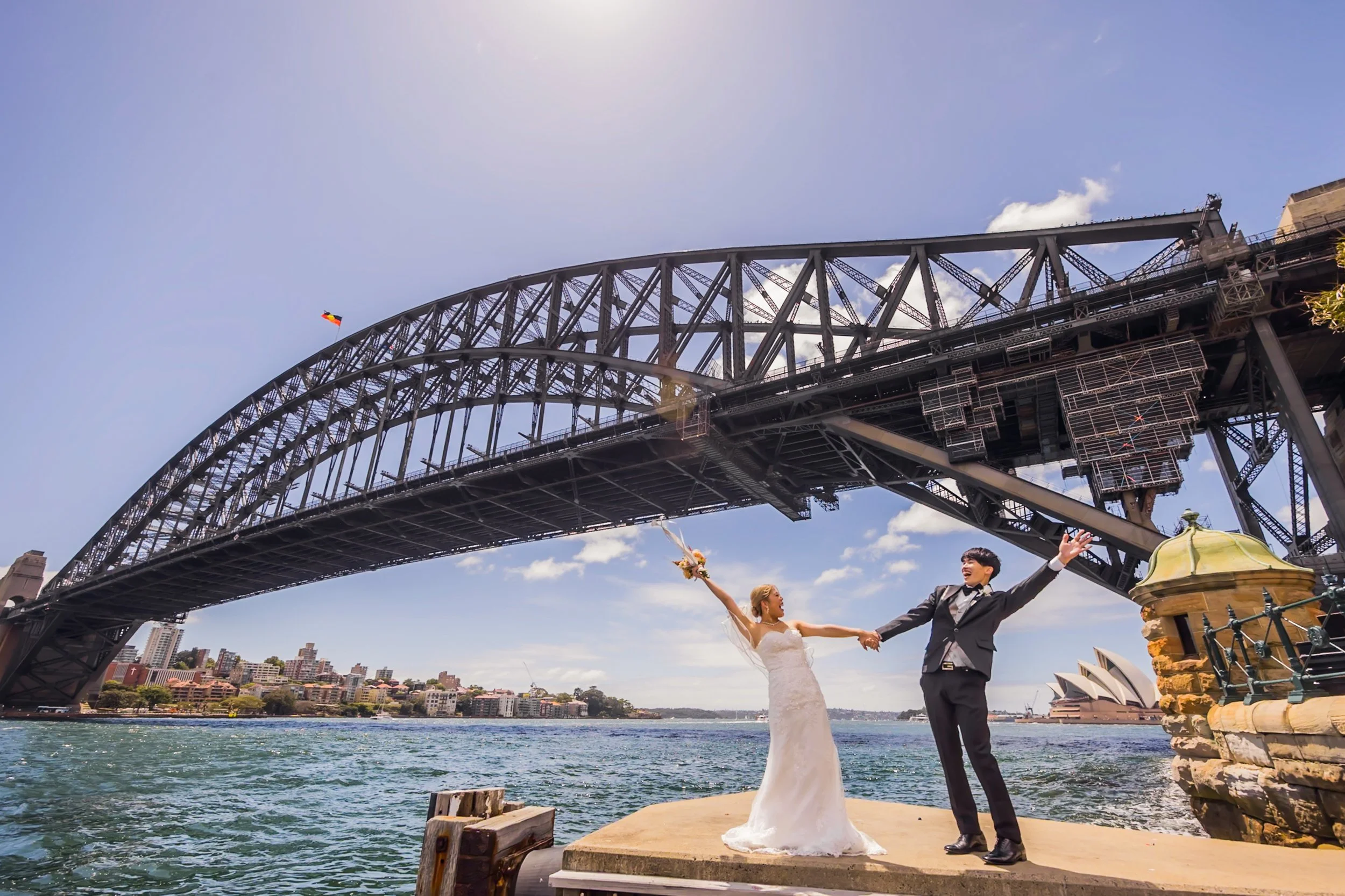 Bride and groom holding hands under the Sydney Harbour Bridge with Sydney Opera House in the background, celebrating their wedding.