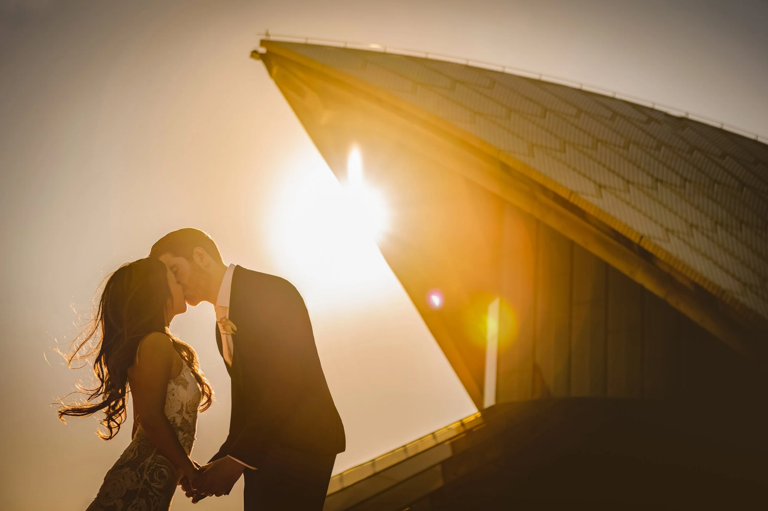 A couple dressed in wedding attire sharing a kiss with the sun shining behind Sydney Opera House