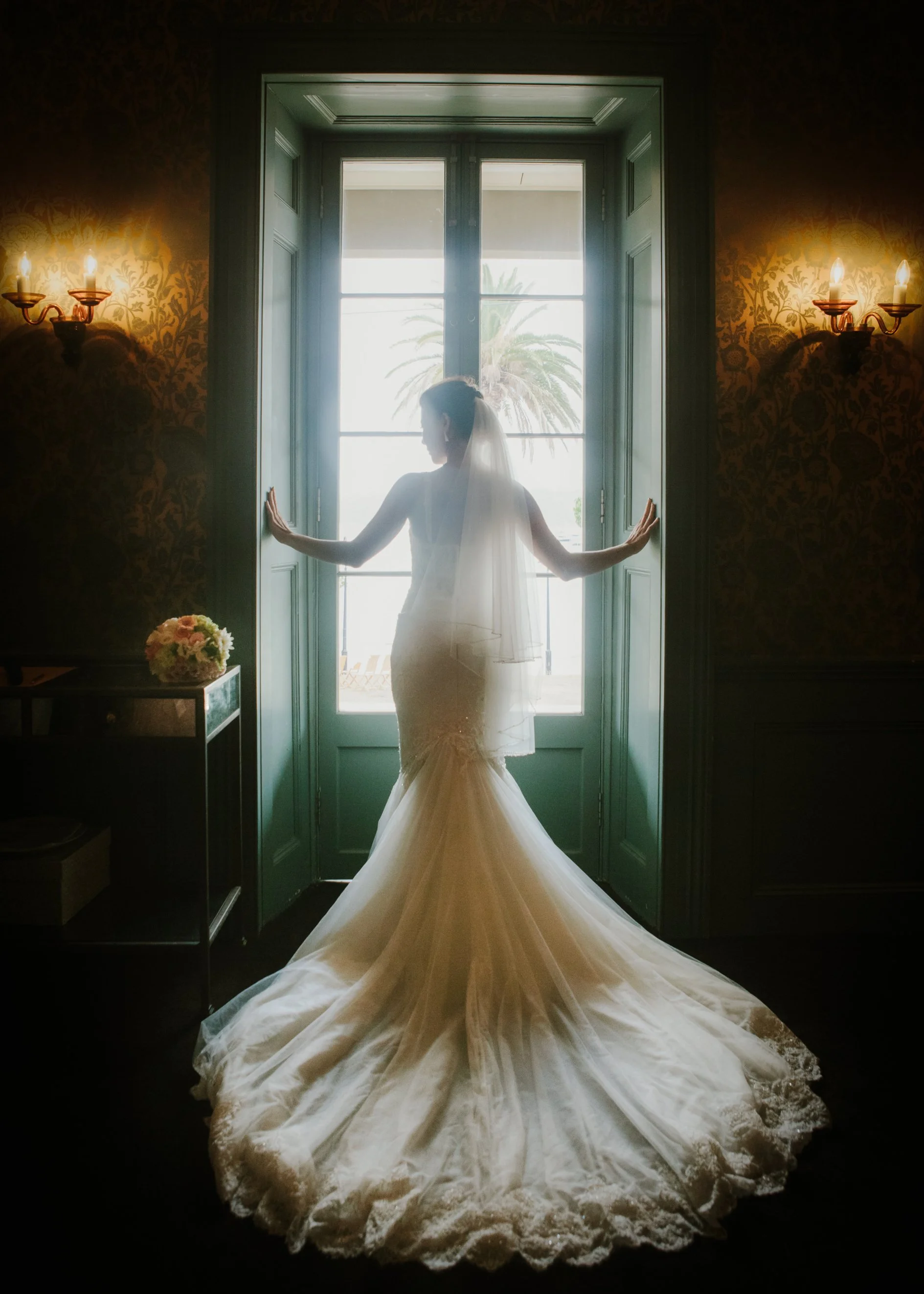 A bride in a wedding gown stands by a large window, looking outside with her hands resting on the window frame. She is silhouetted against the bright light coming through the window, with a palm tree visible outside. The room has ornate wallpaper and