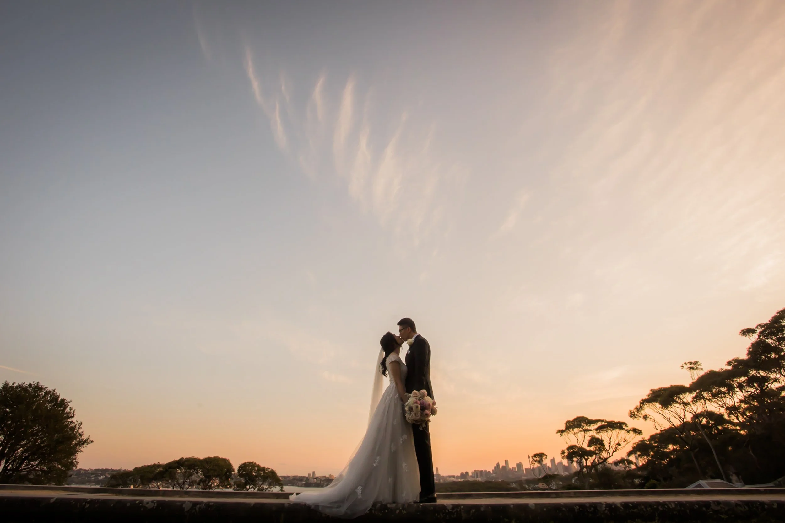 Silhouette of a bride and groom kissing at sunset with city skyline and trees in the background.