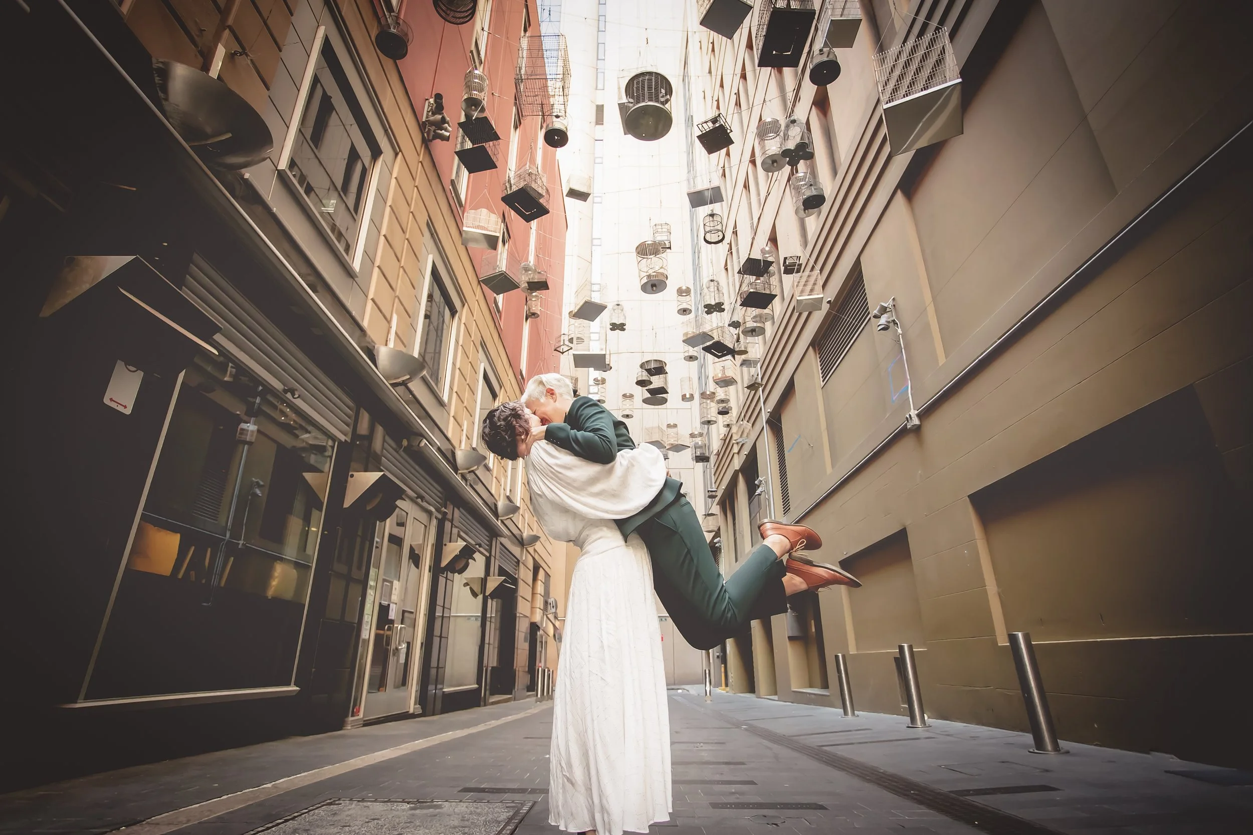 A couple kissing in an alleyway, with the man holding the woman in his arms. The alley is decorated with hanging birdcages and has tall buildings on either side.