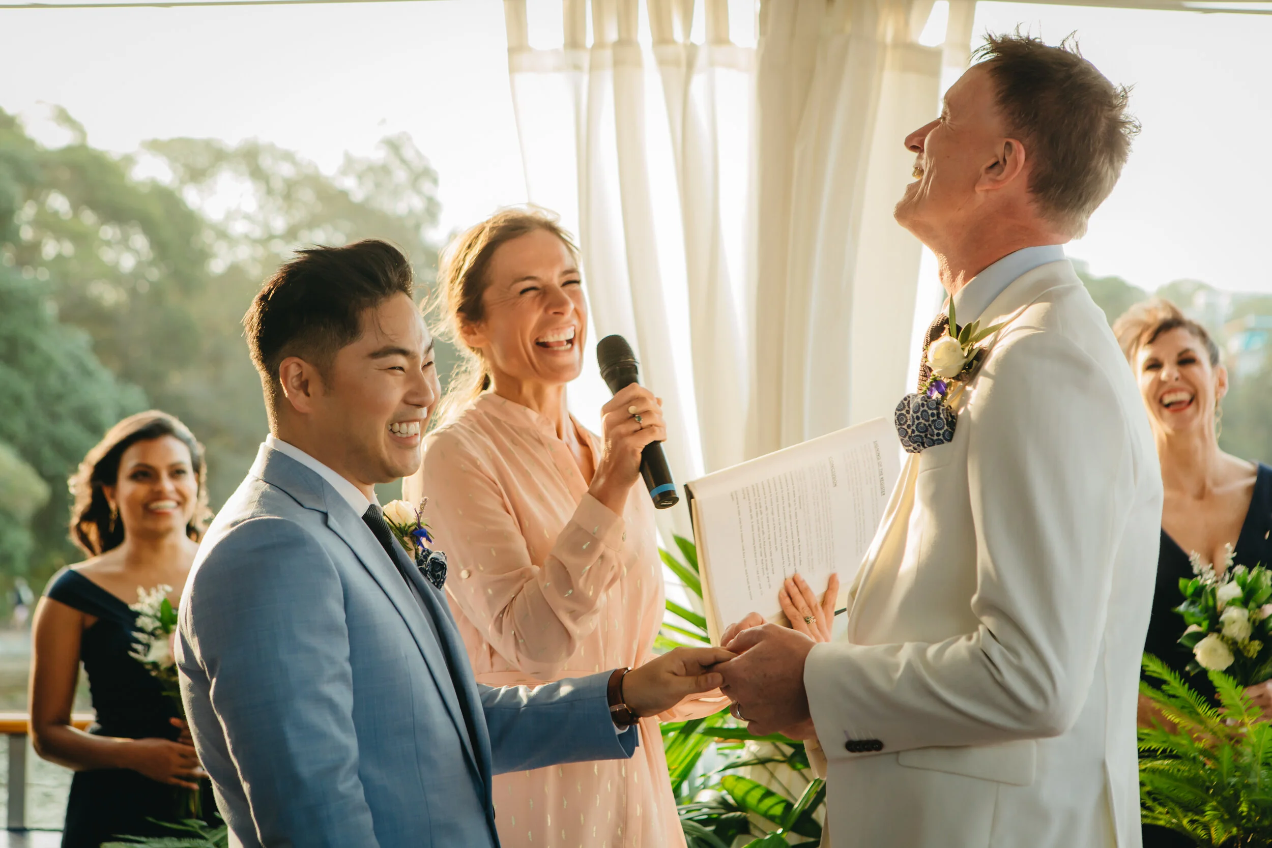 A wedding ceremony with the couple holding hands, the officiant smiling and holding a microphone, and the officiant reading from a book outdoors with friends and family in the background.