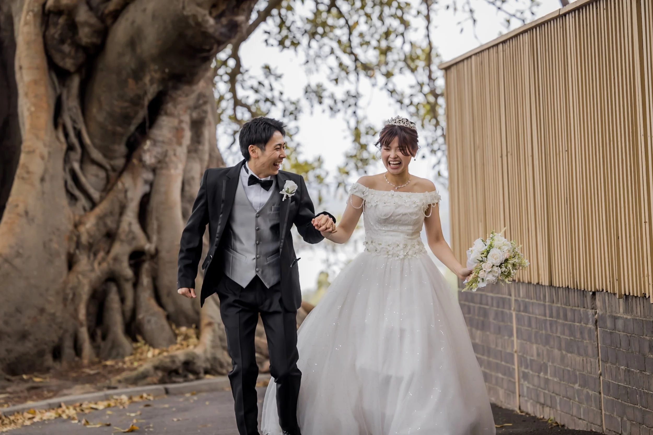 A happy bride and groom walking outdoors, holding hands and smiling, with the bride in a white wedding gown and the groom in a black tuxedo, in front of large tree roots and a wooden fence.