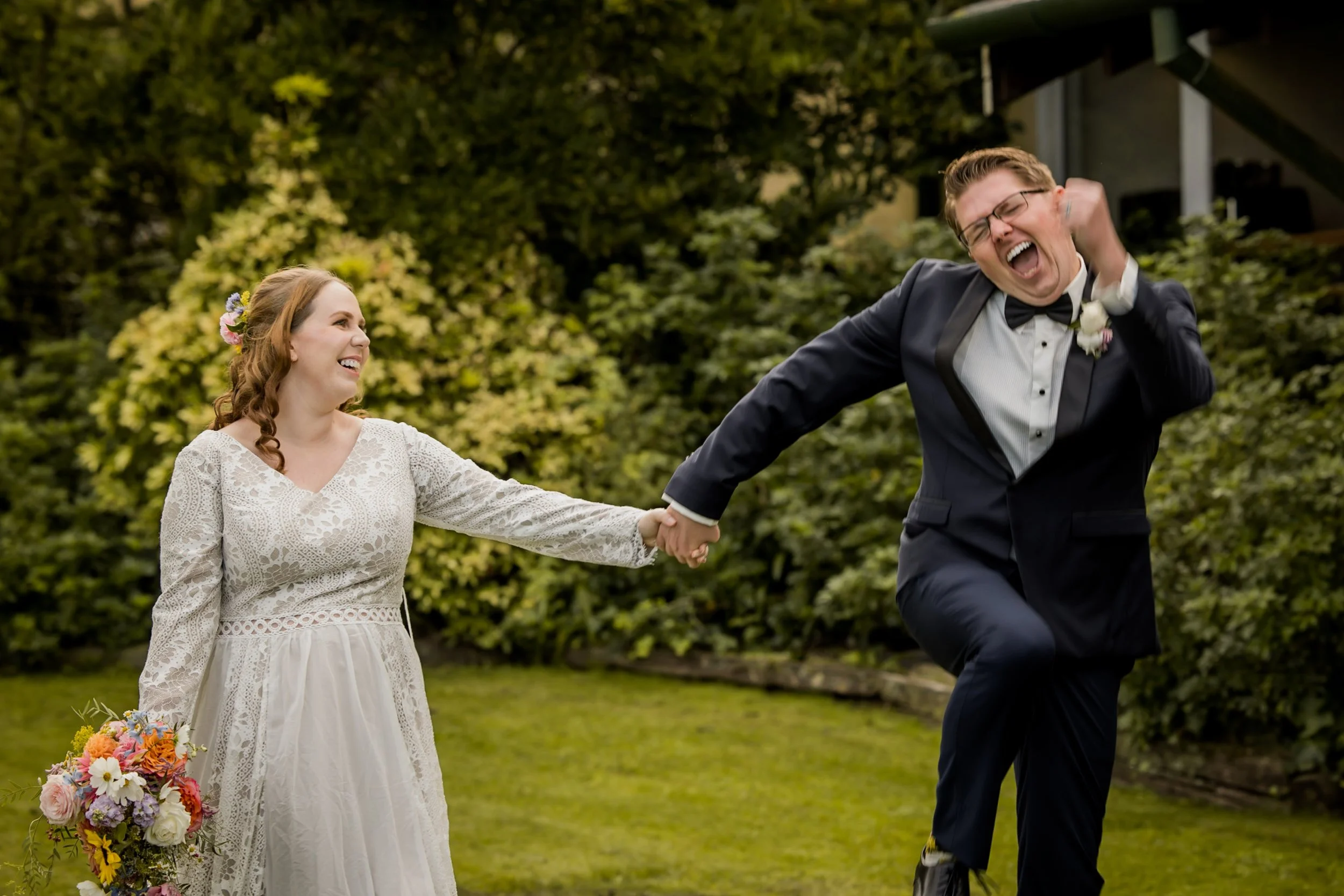 Bride and groom holding hands outside, with the groom lifting his leg and laughing, bride smiling and holding a bouquet of flowers, greenery in the background.