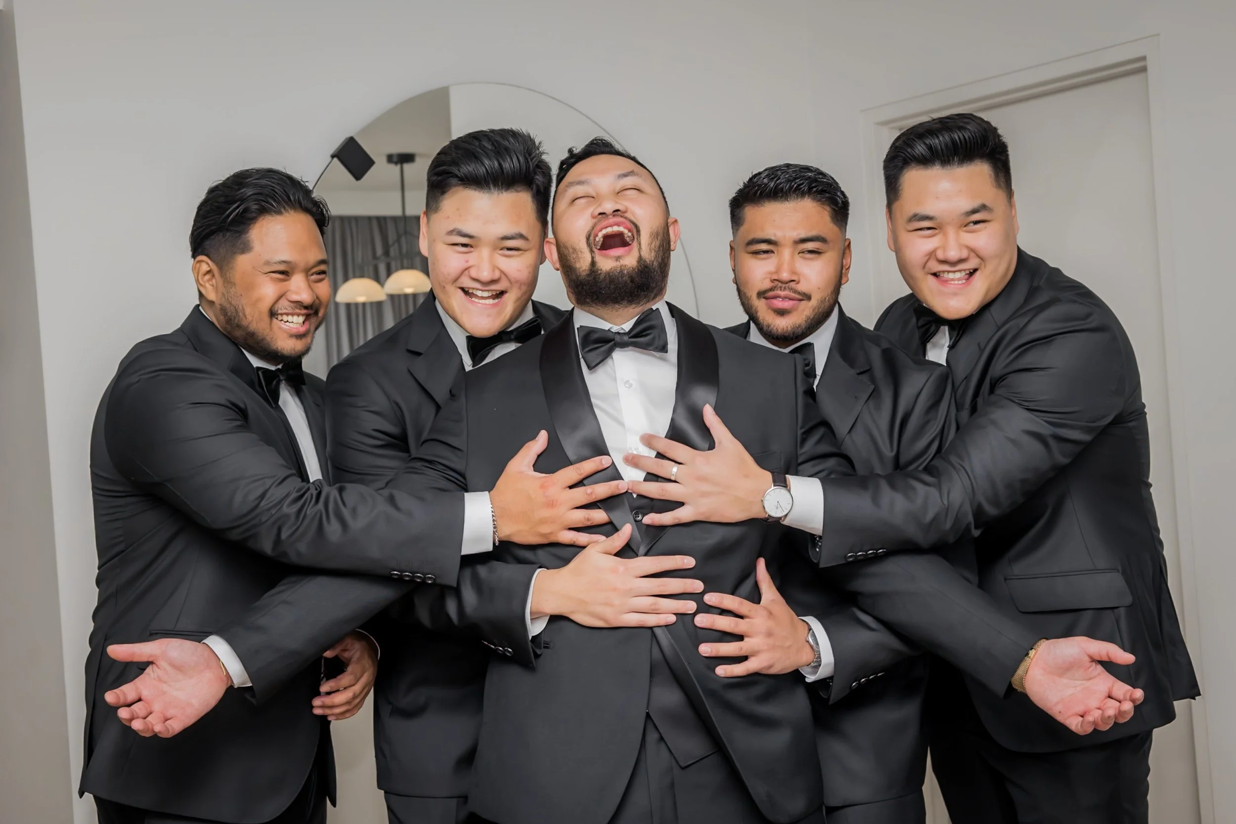 Group of five men in tuxedos celebrating, with one in the center laughing and surrounded by friends who are hugging him.