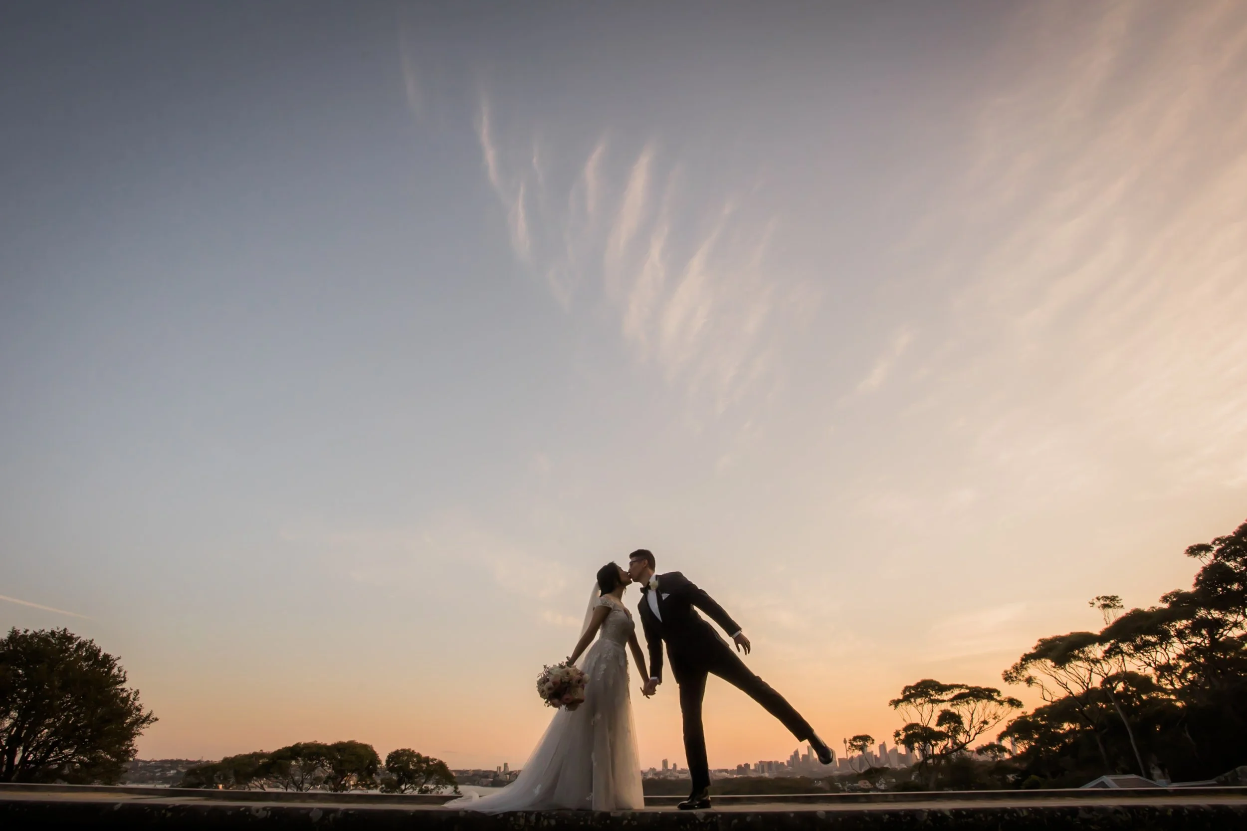 A couple dressed in wedding attire sharing a kiss at sunset, with the groom leaning slightly to kiss the bride, holding her hands, holding a bouquet of flowers, with a city skyline and trees in the background at Gunners Barracks, Mosman