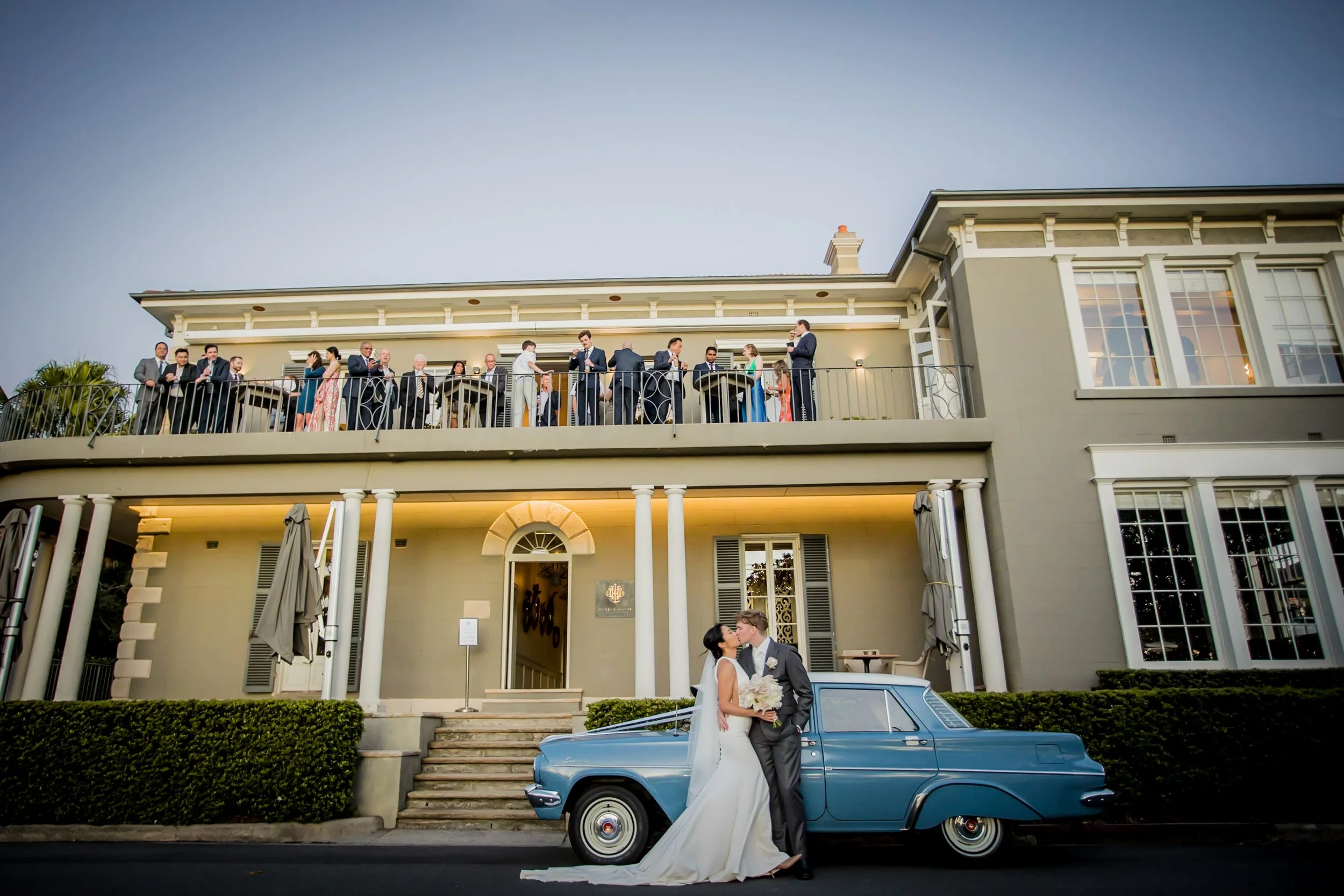 A newlywed couple kissing in front of a classic blue car outside a large, multi-story house during their wedding celebration, with guests mingling on the upper balcony.