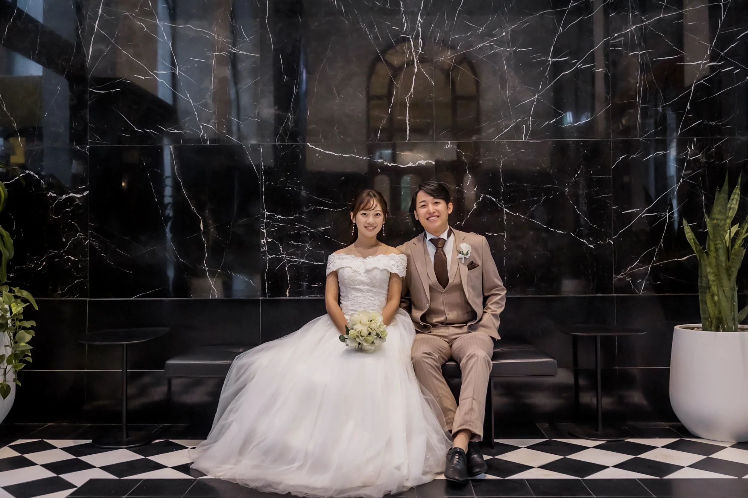 A bride and groom sitting on a black bench in front of a black marble wall, smiling for a wedding photo. The bride is wearing a white wedding gown and holding a bouquet, and the groom is dressed in a beige suit with a brown tie.