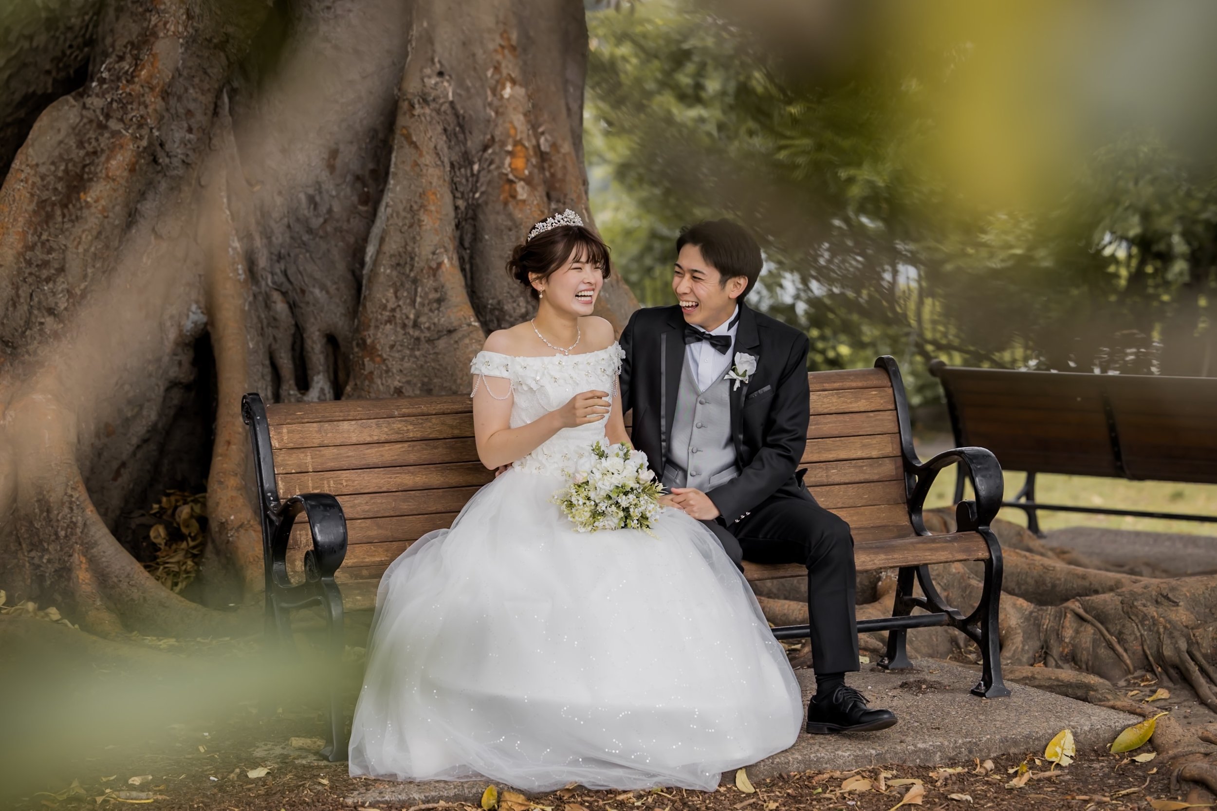 Wedding couple seated on a park bench laughing, with large tree trunk behind them and green foliage around.