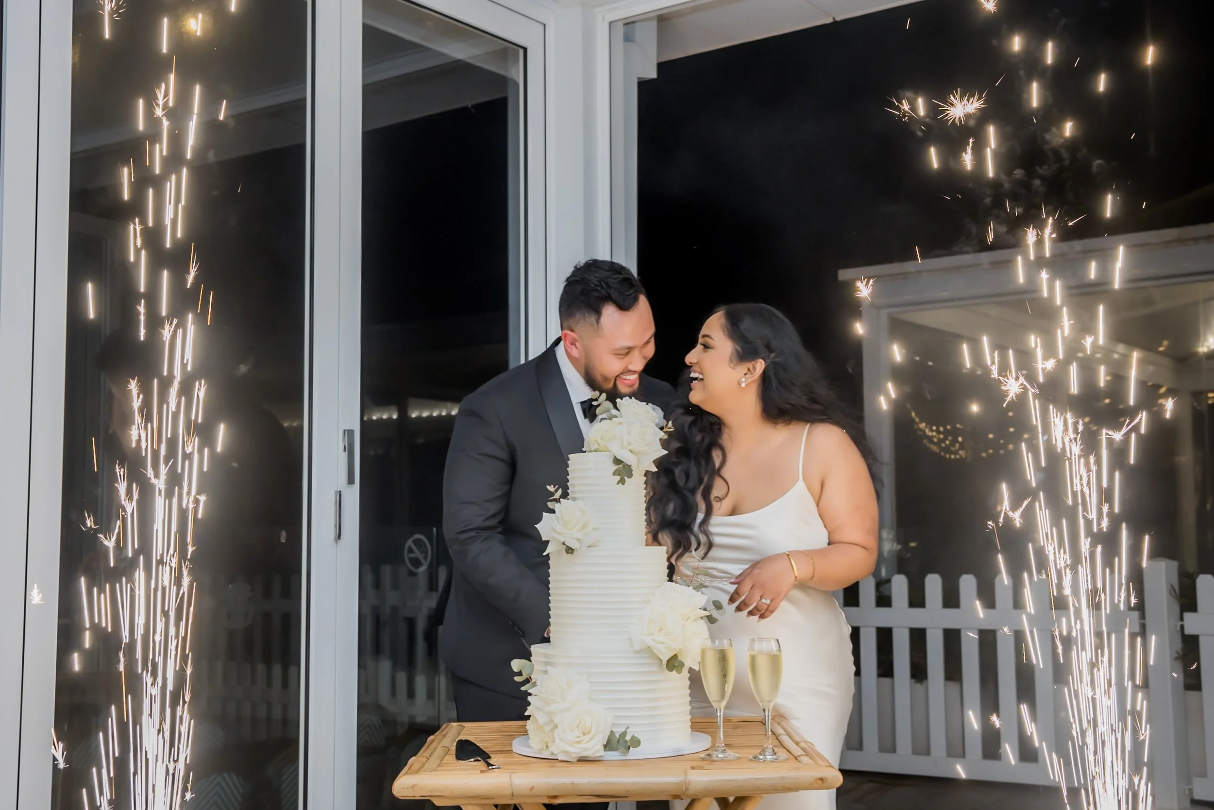 A newlywed couple is celebrating their wedding with a cake-cutting ceremony. They are smiling at each other near a tall white wedding cake decorated with white roses, with two glasses of champagne on a small wooden table.  Long Reef Golf Course