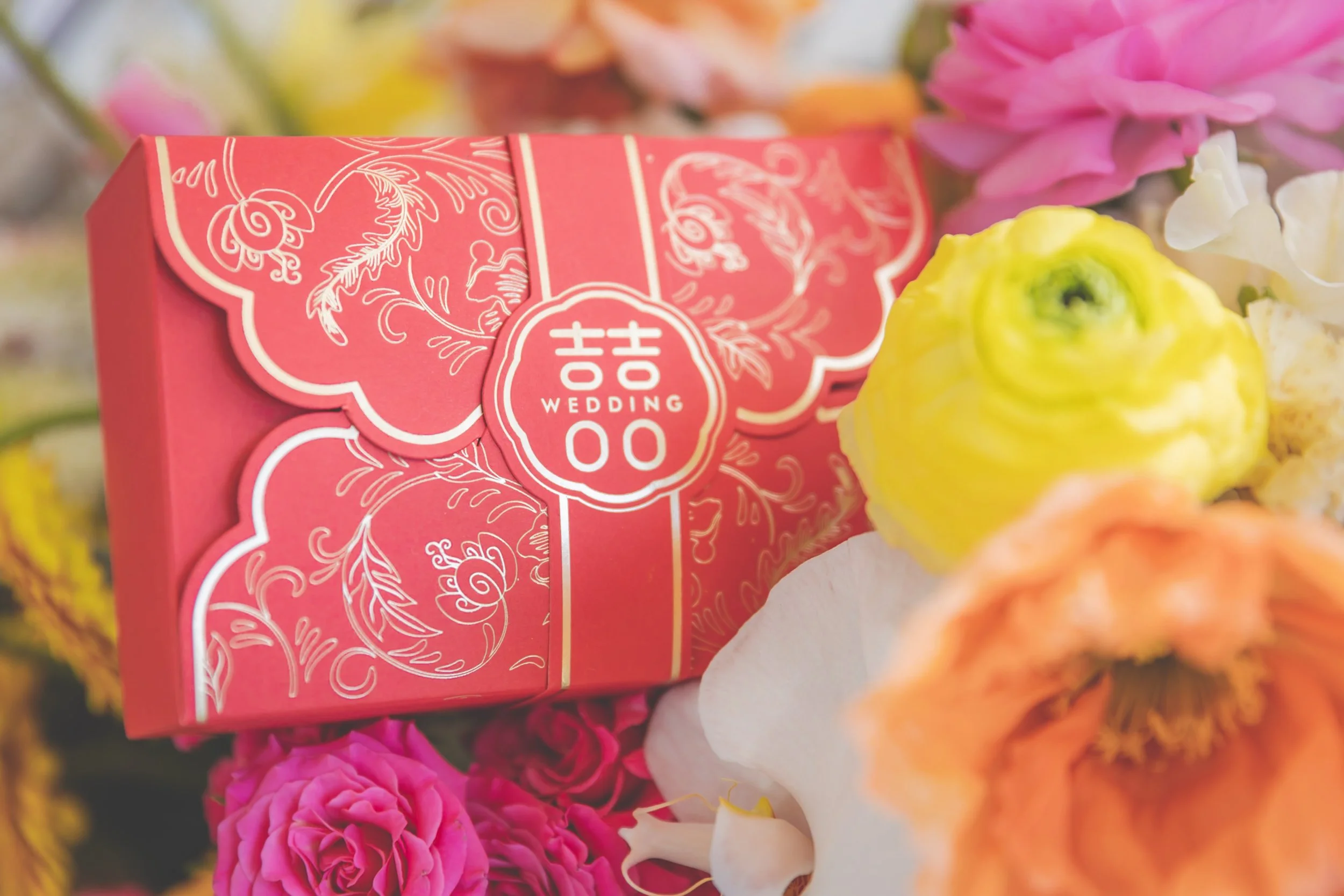 A red wedding favor box with white intricate floral designs and the words 'Wedding Gift' on the top, surrounded by colorful flowers, including pink, yellow, white, and peach blooms.