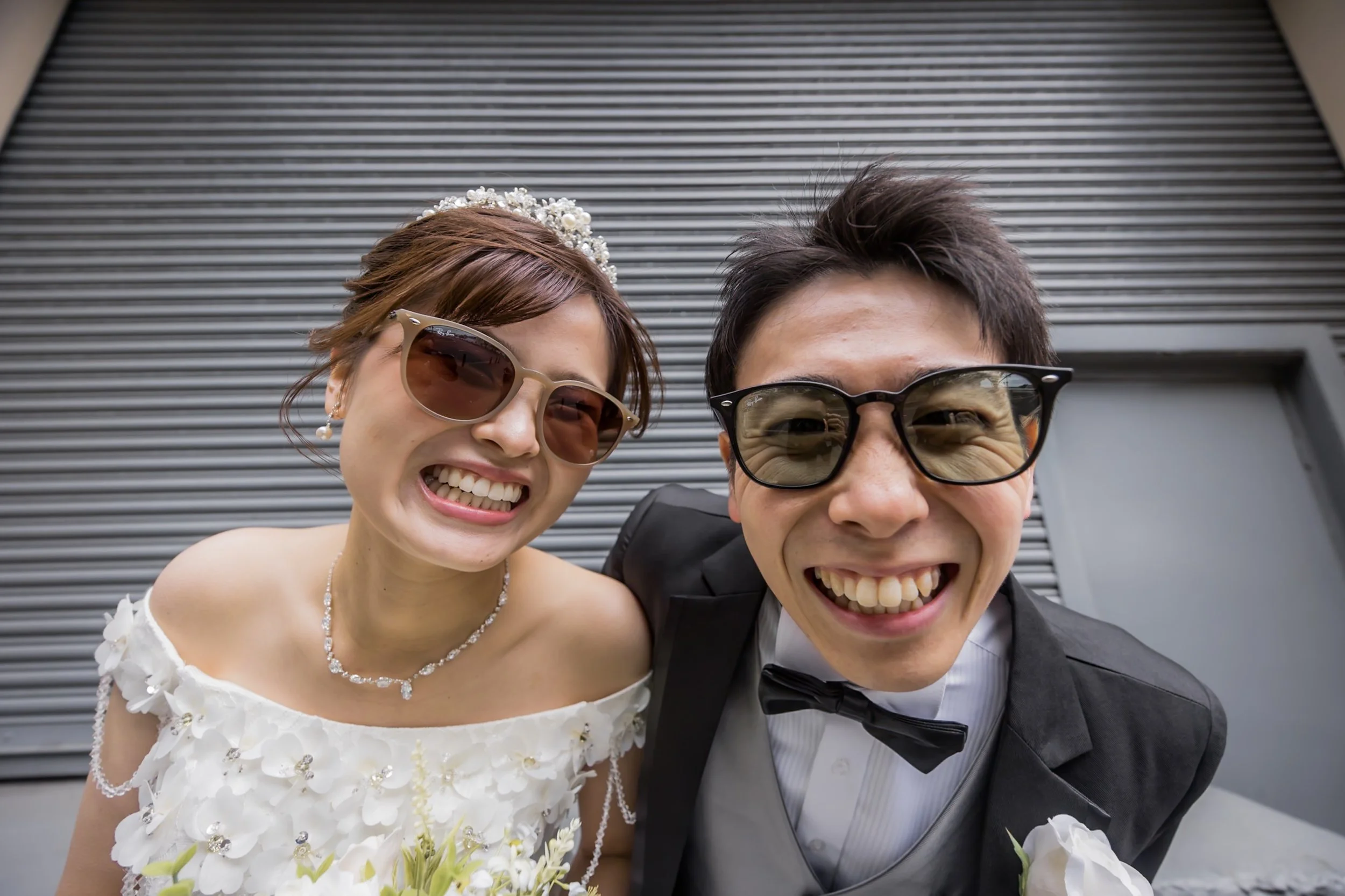 A smiling couple dressed in wedding attire, wearing sunglasses and leaning close together in front of a metal shutter.