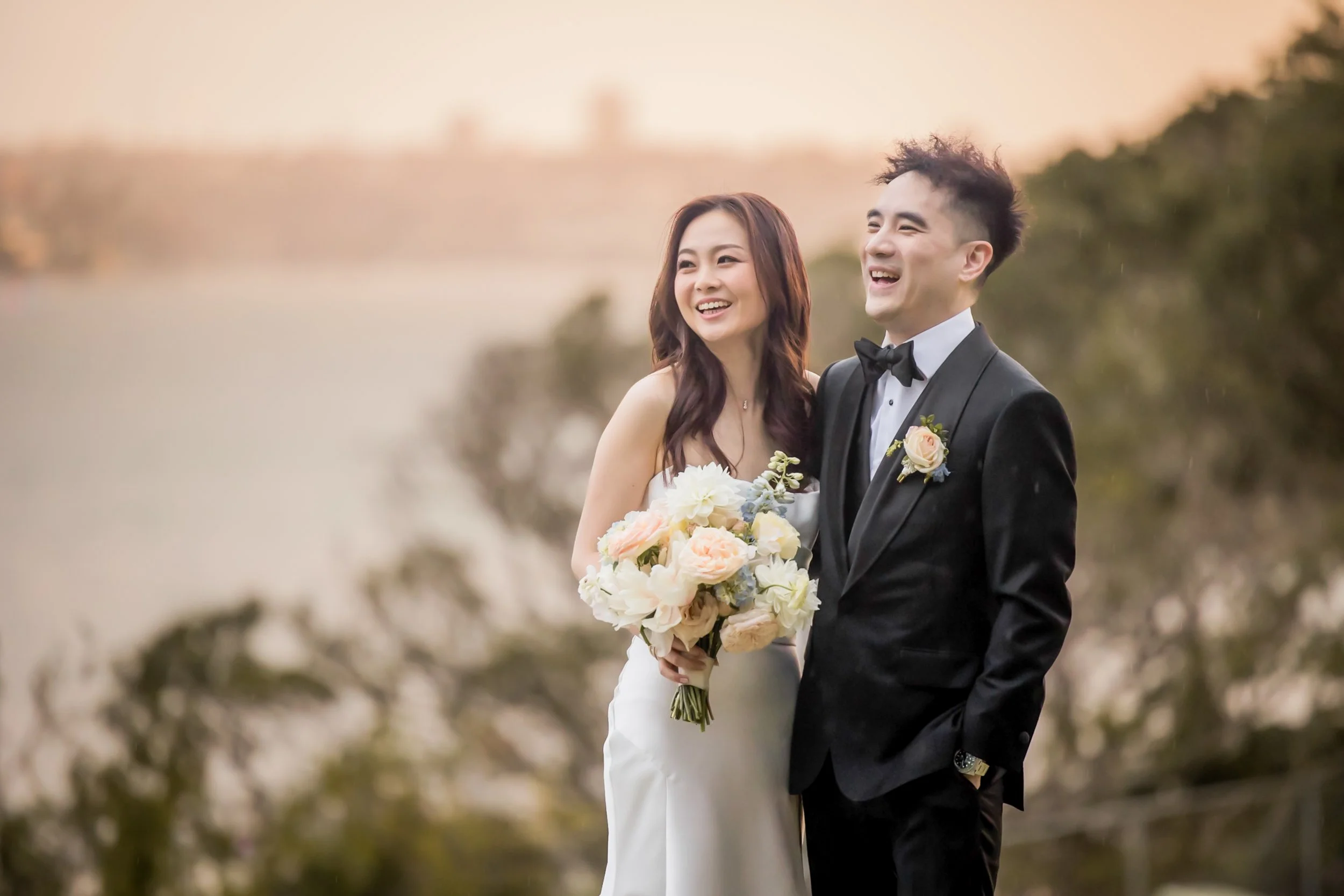 A happy bride and groom standing outdoors during their wedding, with the bride holding a bouquet of flowers and both dressed in formal attire, smiling at each other.  Sergeants Mess Sydney - Katsu Nojiri Sydney Wedding Photographer
