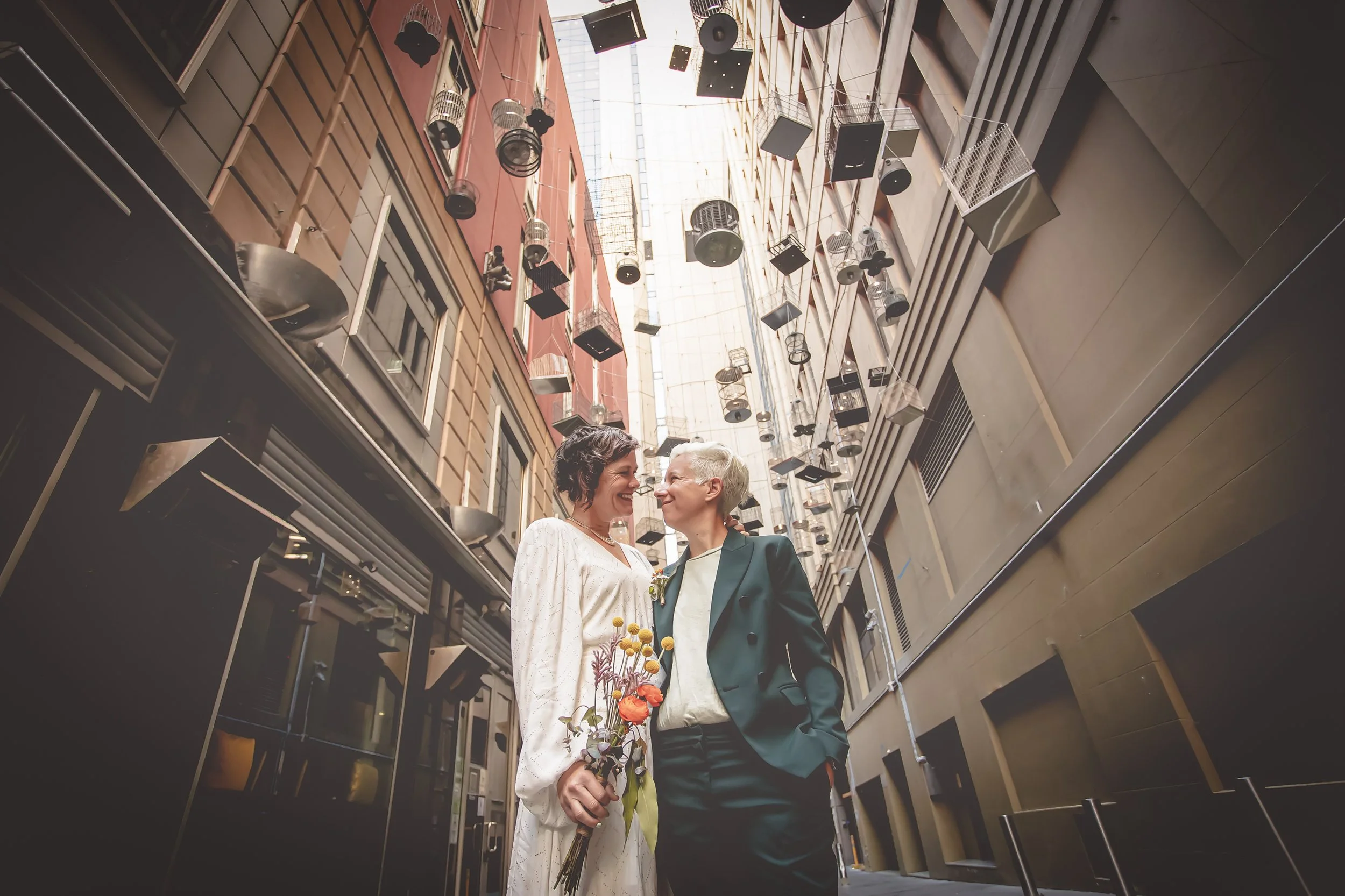 Two women standing close together in an alley with hanging lights and air conditioning units, smiling and holding a bouquet of flowers.