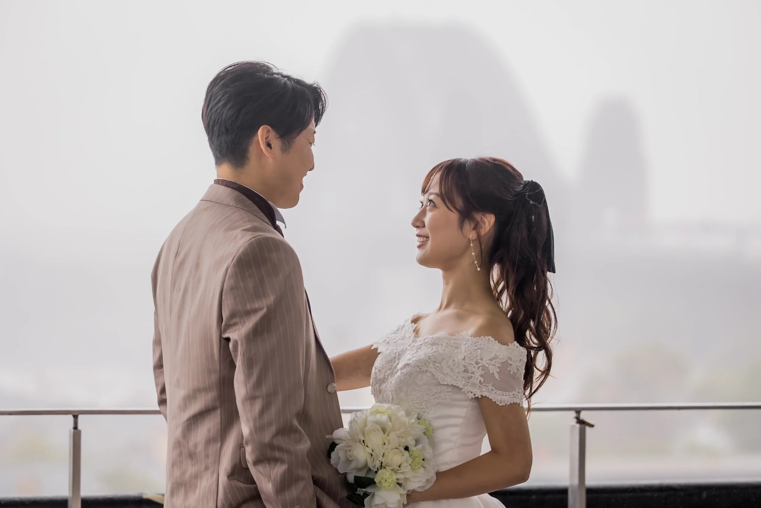 A bride and groom standing close on a balcony, gazing into each other's eyes with buildings in the background.