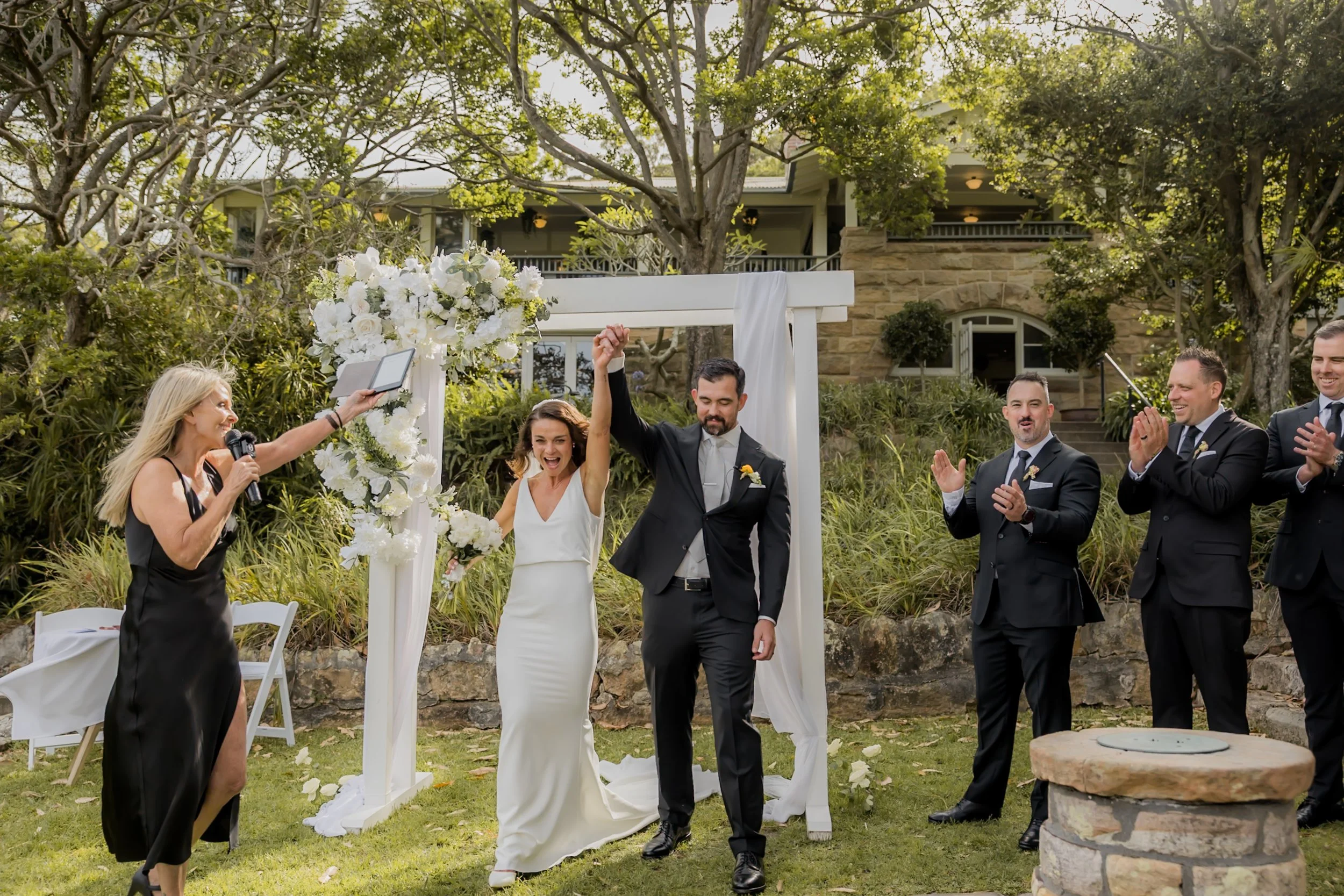 A newlywed couple at their outdoor wedding ceremony, the bride and groom holding hands and smiling, with guests clapping and a woman officiant speaking into a microphone, all under a floral arch decorated with white flowers, surrounded by trees and a