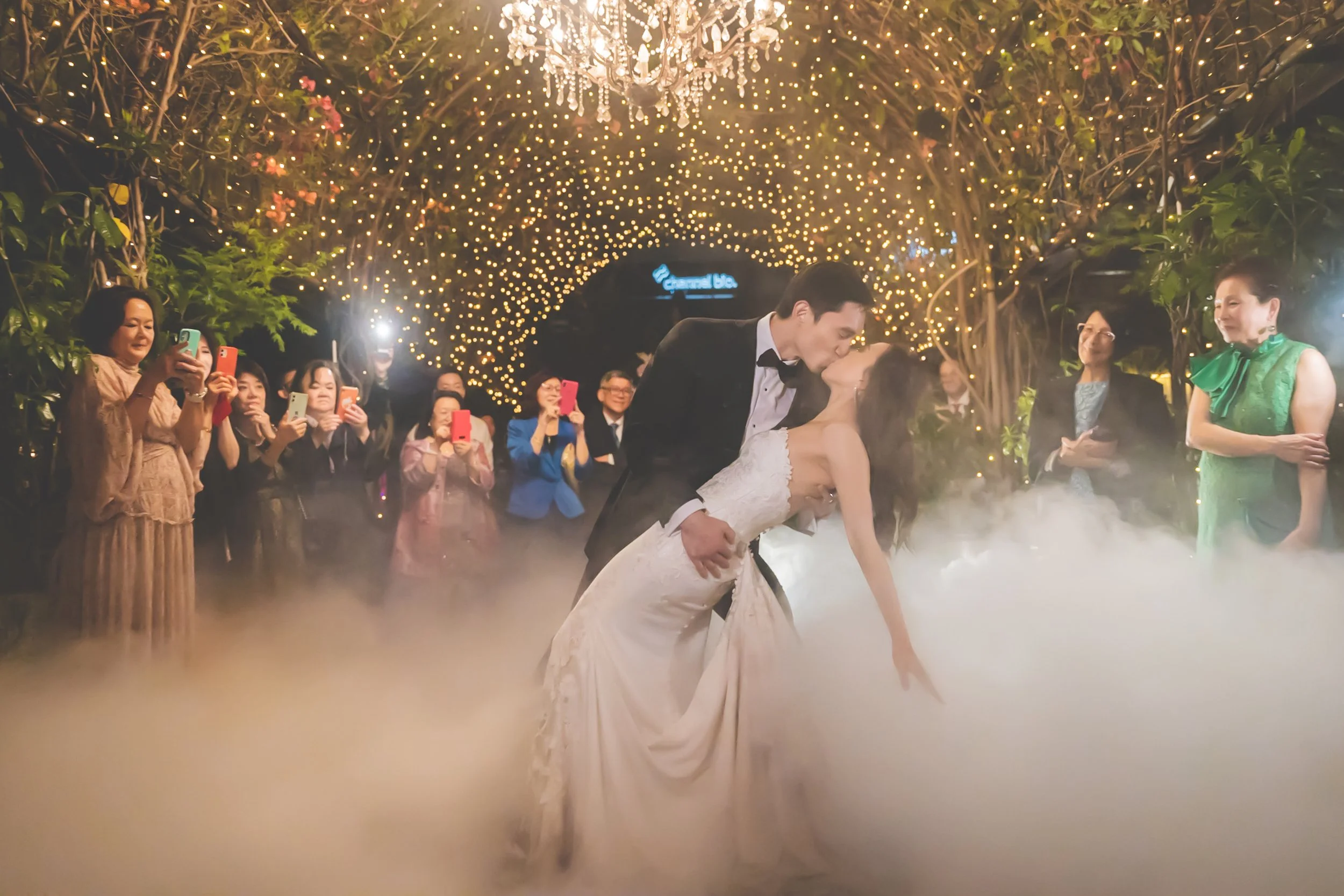 A newlywed couple sharing a kiss during their wedding dance under a canopy of hanging string lights, with guests watching and taking photos, and fog on the dance floor.