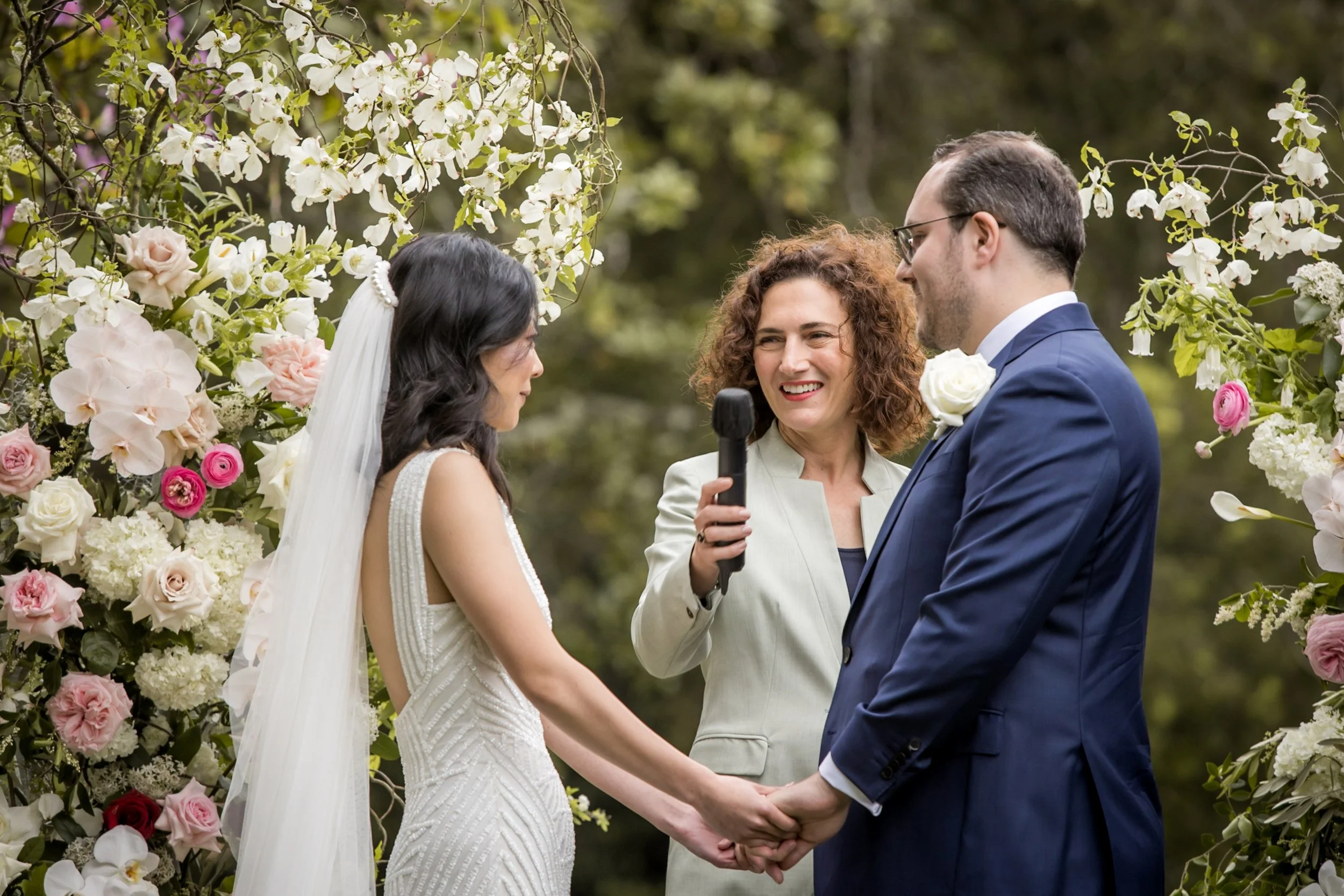 A wedding ceremony outdoors with a bride and groom holding hands under a floral arch as an officiant officiates, with the couple gazing at each other and smiling.