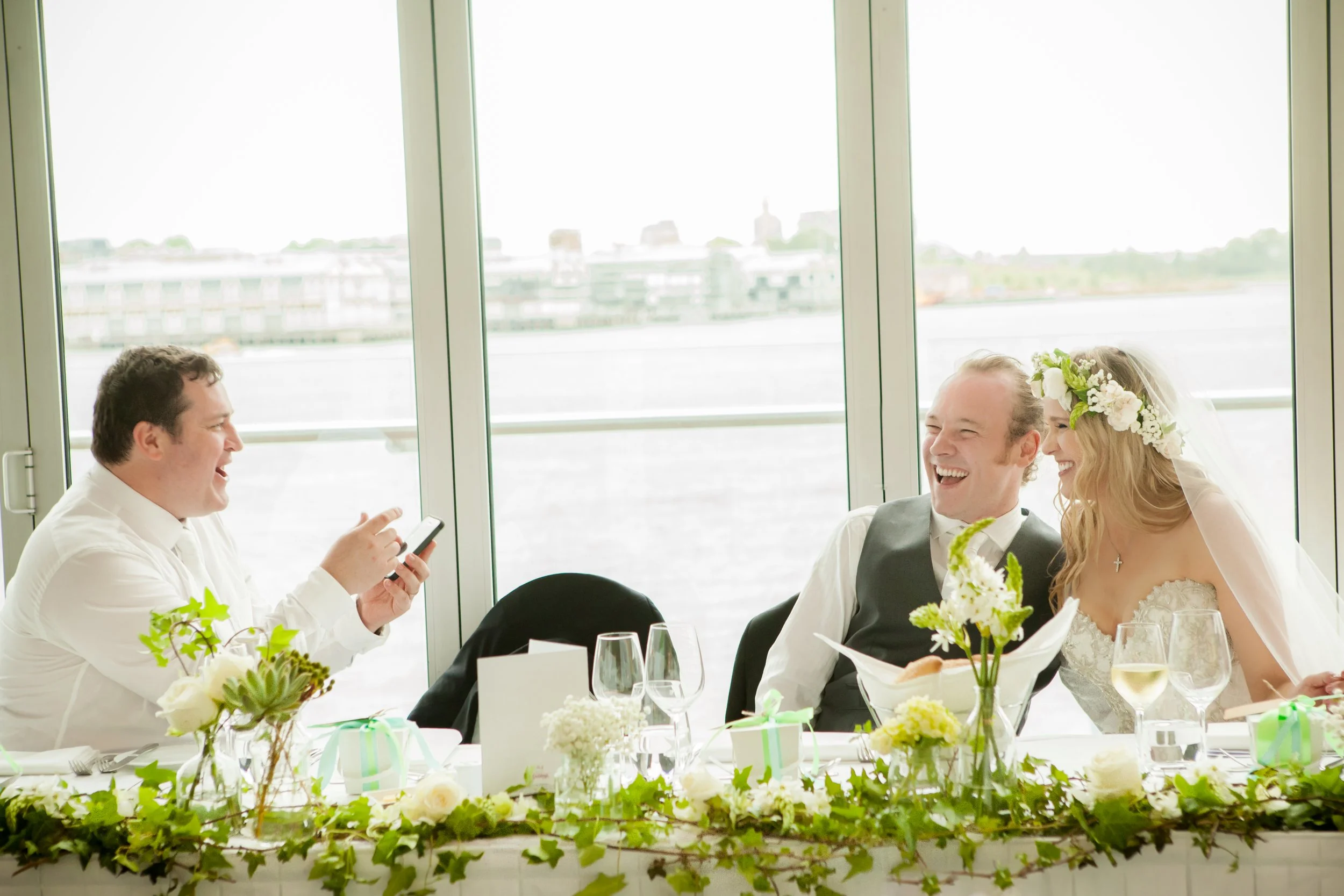 A wedding reception with a bride, groom, and another man sitting at a decorated table, laughing and taking a photo, with large windows overlooking a body of water in the background.
