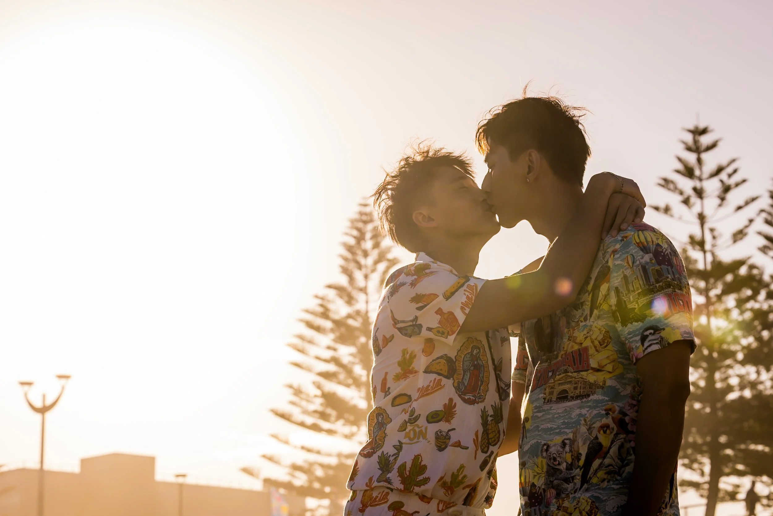 Two men kissing outdoors during sunset, with trees and buildings in the background.