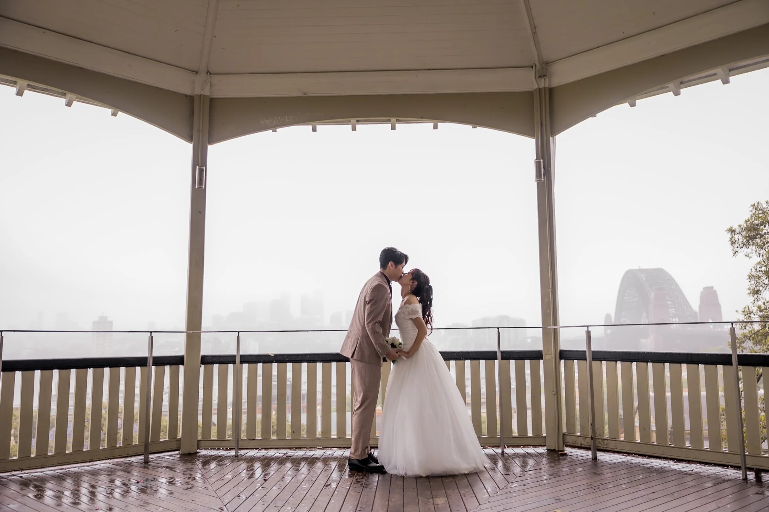A bride and groom kiss on a covered balcony with city skyline in the background, wet wooden floor, and foggy weather.