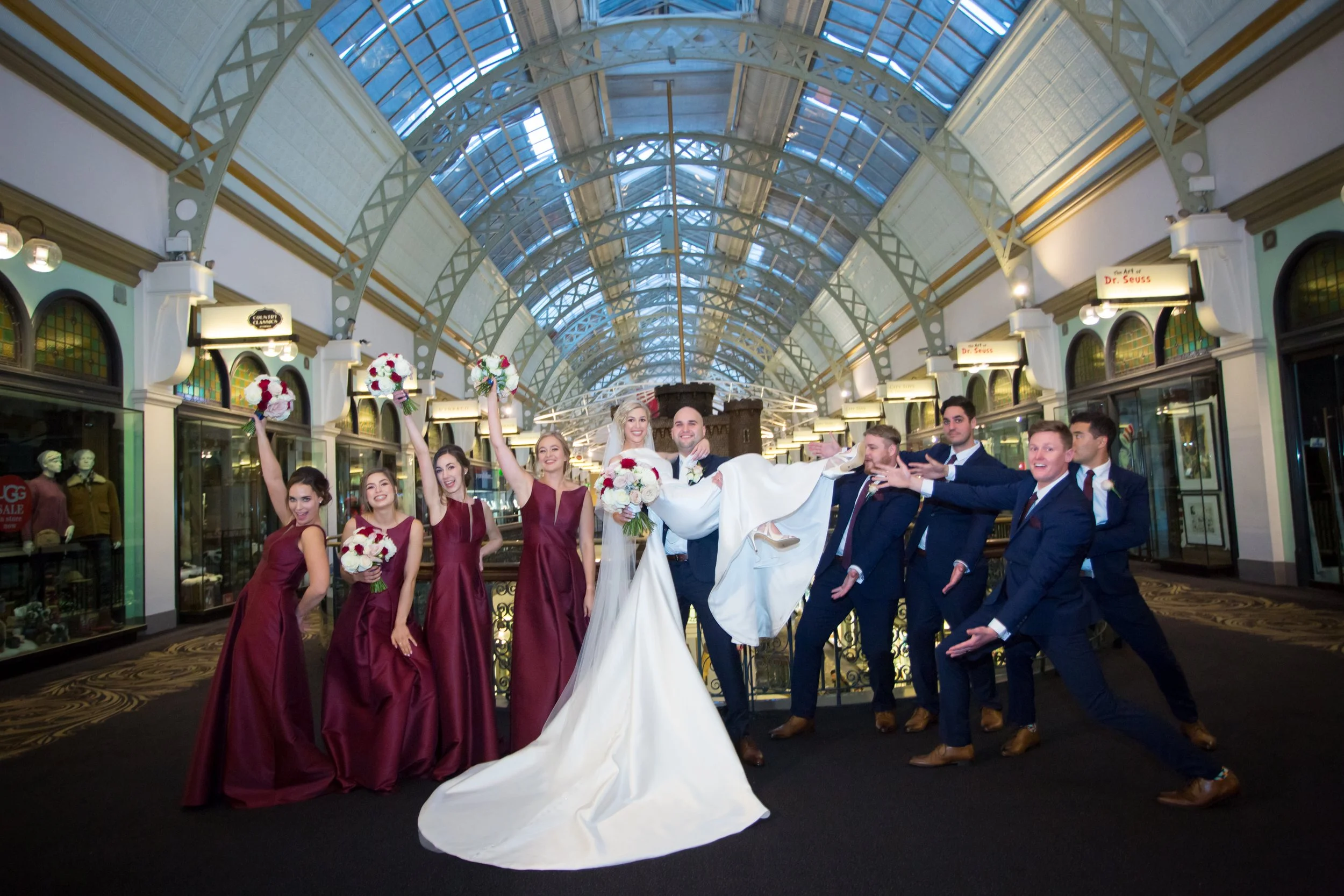 A wedding party inside a train station with high arched glass ceiling. The bride and groom are in the center, with the groom lifting the bride and all members holding bouquets and posing cheerfully. The Tea Room Sydney - Katsu Nojiri Sydney Wedding 