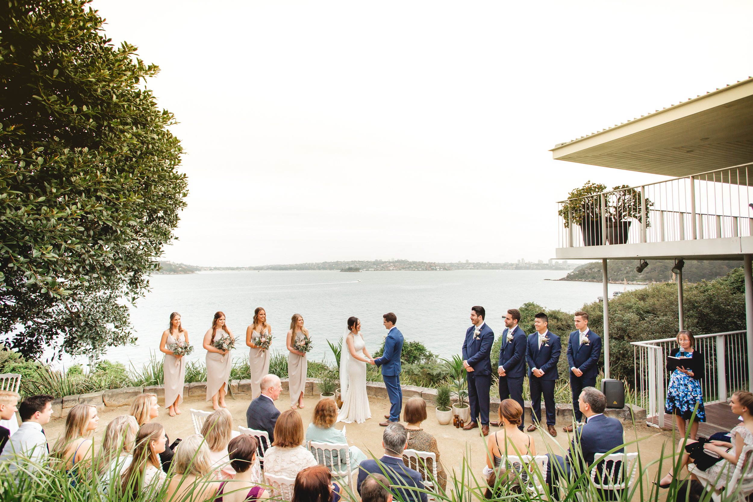 A wedding ceremony taking place outdoors near a body of water, with the bride and groom holding hands and facing each other, surrounded by bridesmaids and groomsmen, with seated guests watching. Sergeants Mess Sydney - Katsu Nojiri Sydney Wedding