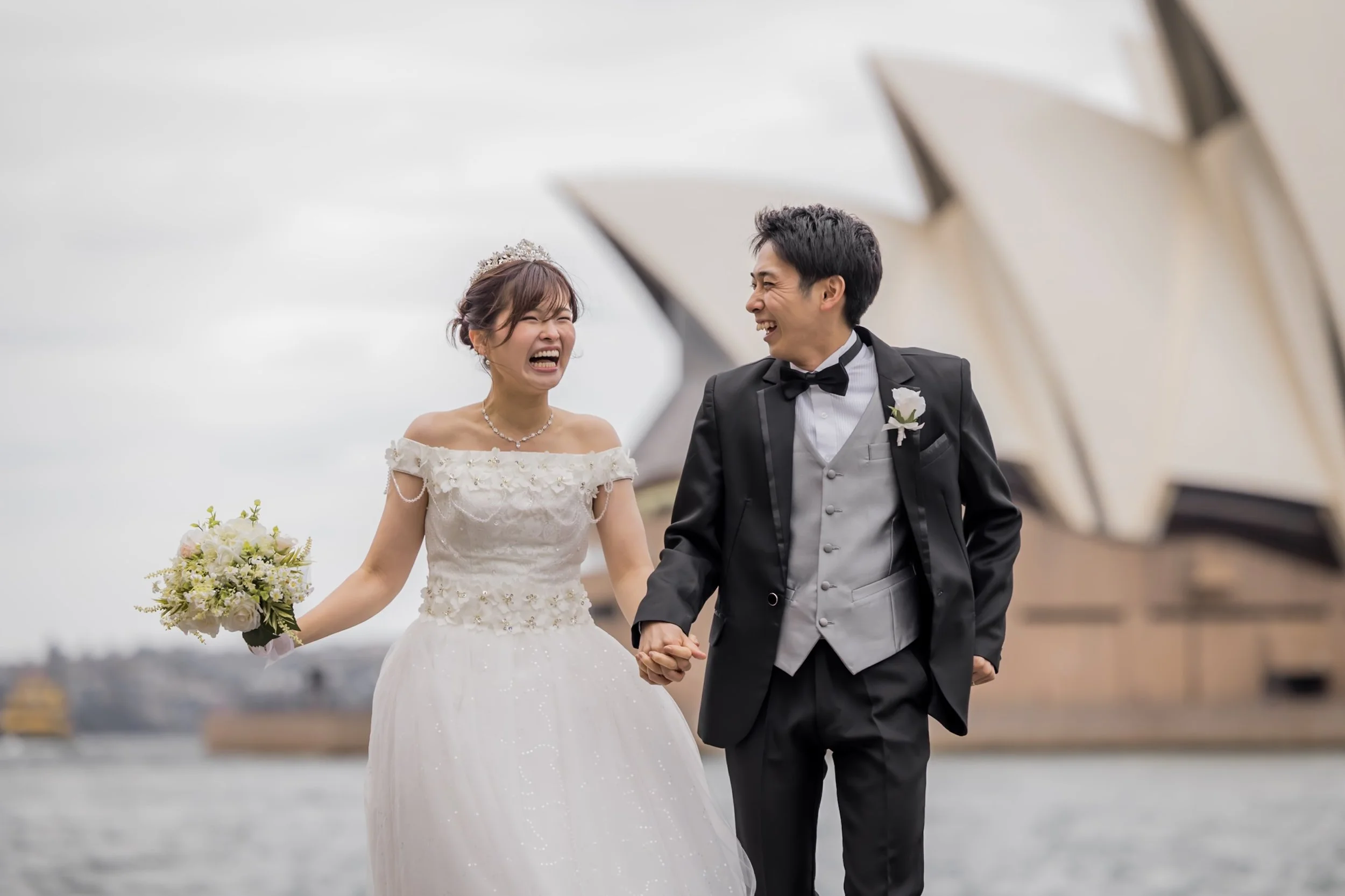 A bride and groom holding hands and laughing in front of the Sydney Opera House.