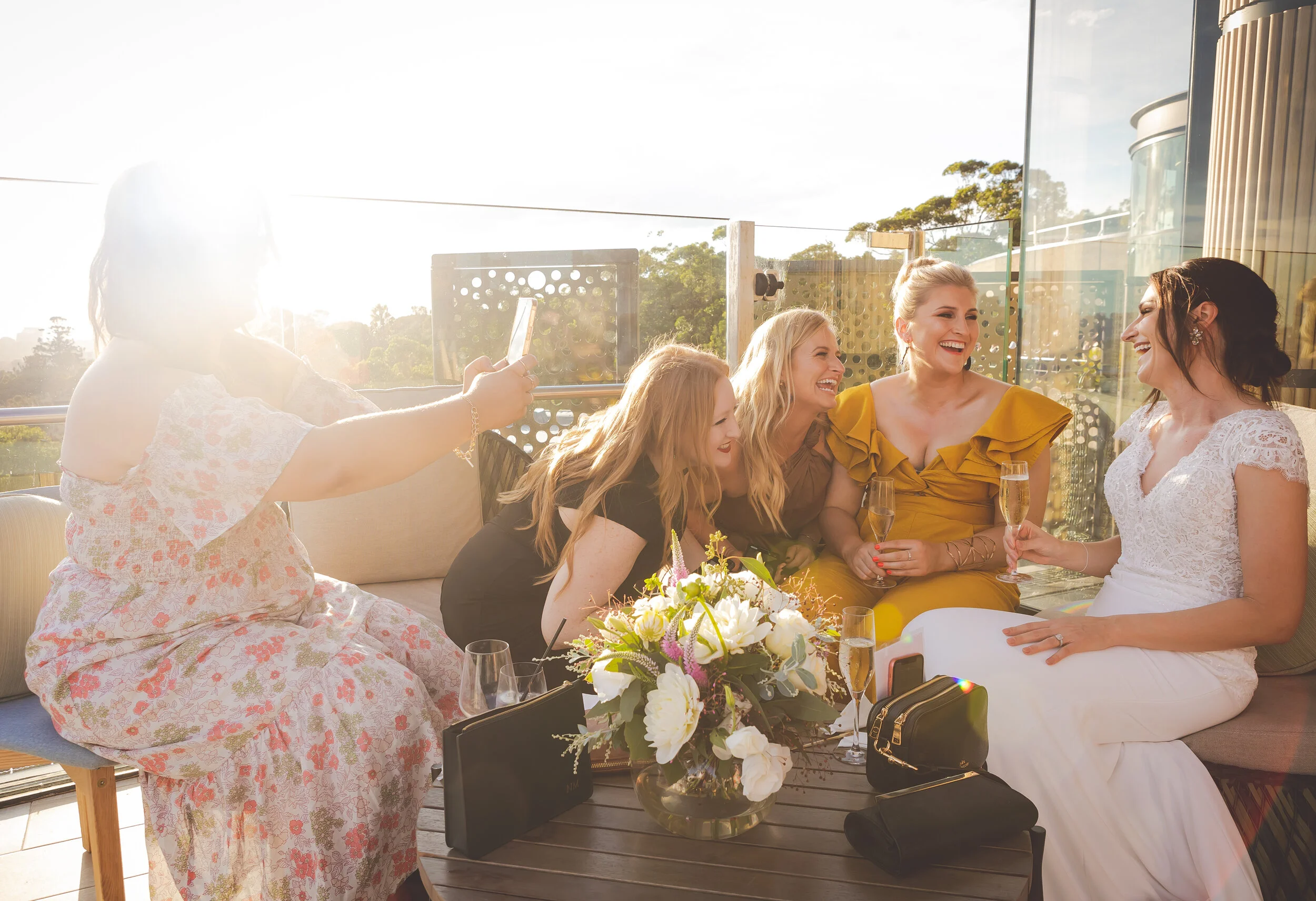 Five women enjoying a gathering on a balcony, with one woman taking a photo, and champagne glasses and a floral centerpiece on the table.